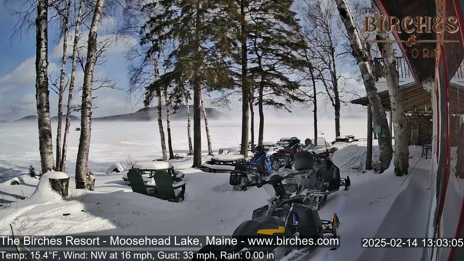 Snow-covered ground with snowmobiles parked near a lakeside resort on a partly sunny, windy day with a misty lake in the background.