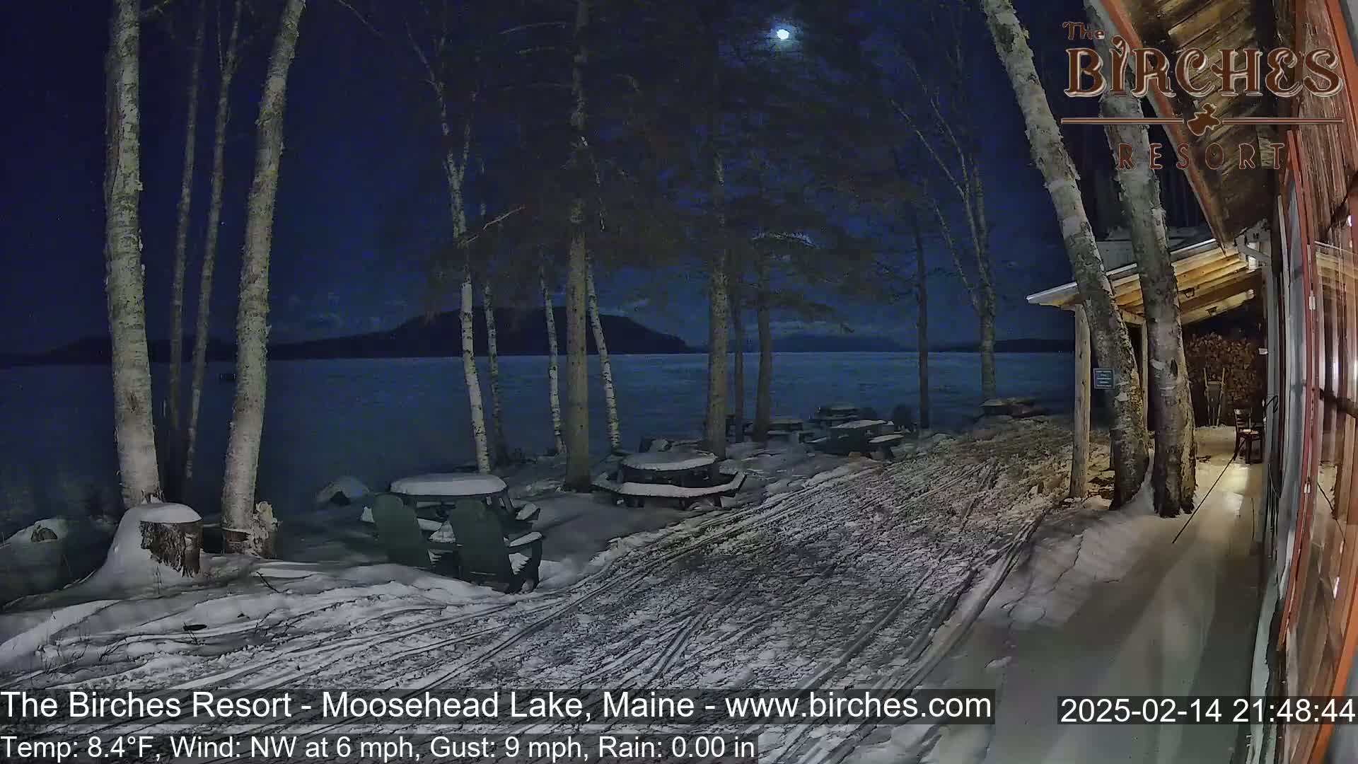 Snow-covered picnic tables sit outside a lakeside building under a nighttime sky with a visible moon.