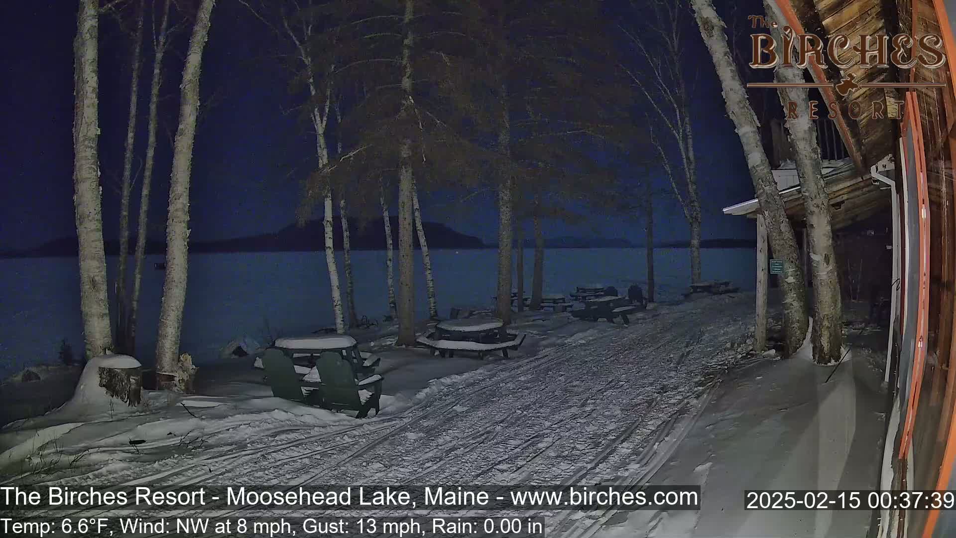 A snow-covered lakeside patio with picnic tables and Adirondack chairs sits under a dark night sky next to a building, with a calm lake visible in the background.