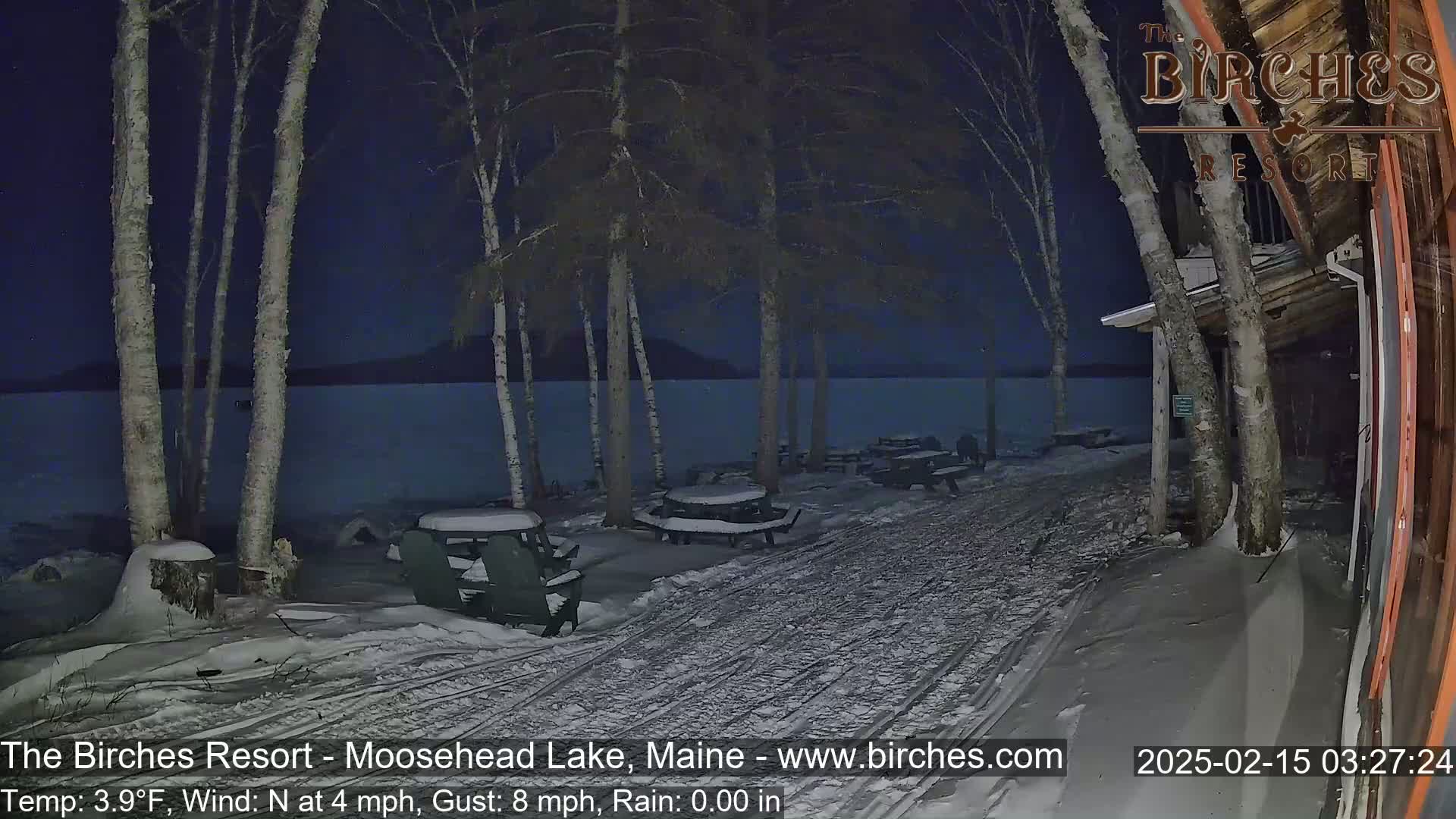 A snow-covered lakeside area at night shows picnic tables and Adirondack chairs near a building under a dark, clear sky.
