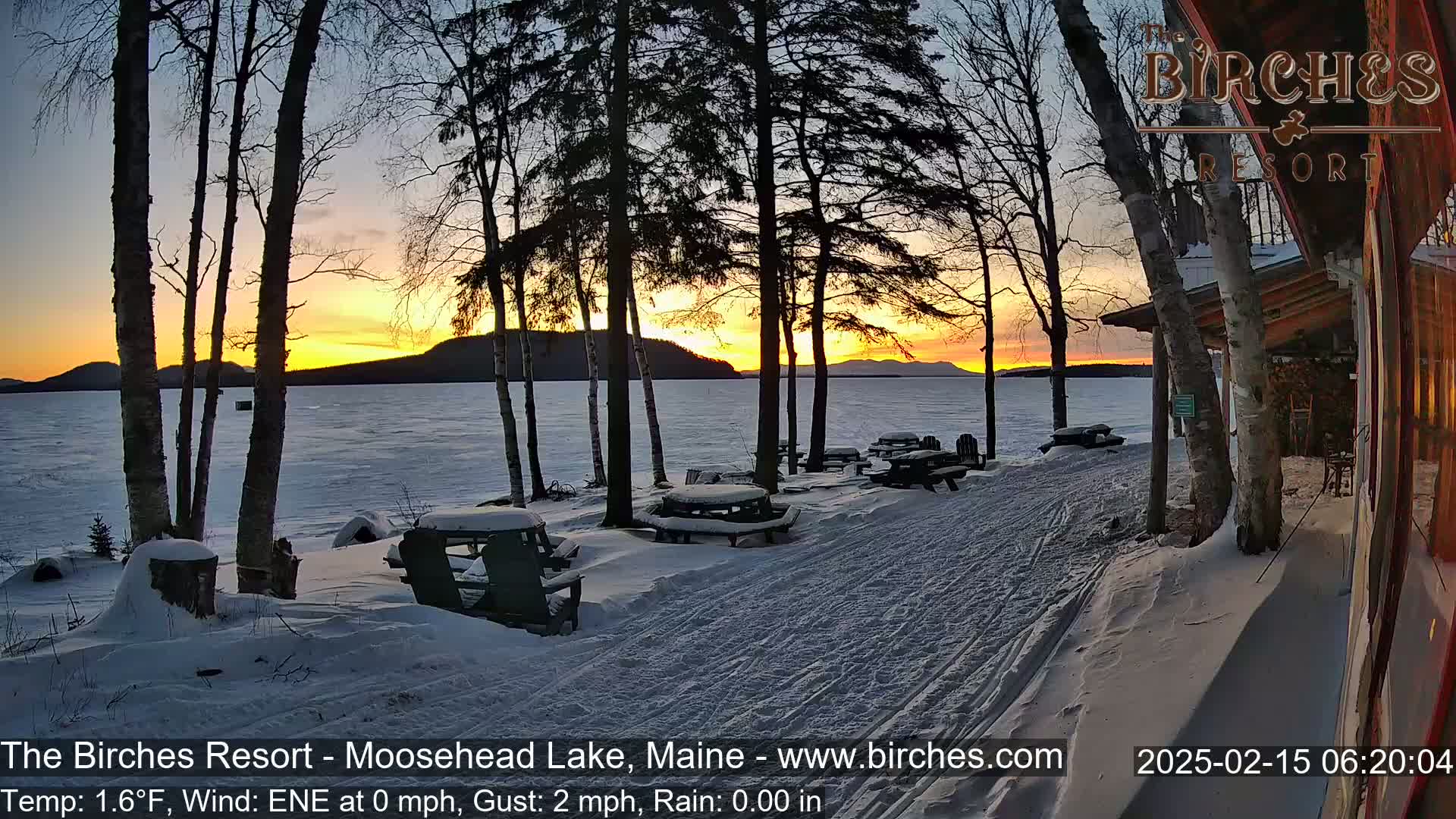 A snowy lakeside scene at sunset shows snow-covered picnic tables and chairs near a building, with a frozen lake and distant mountains in the background.