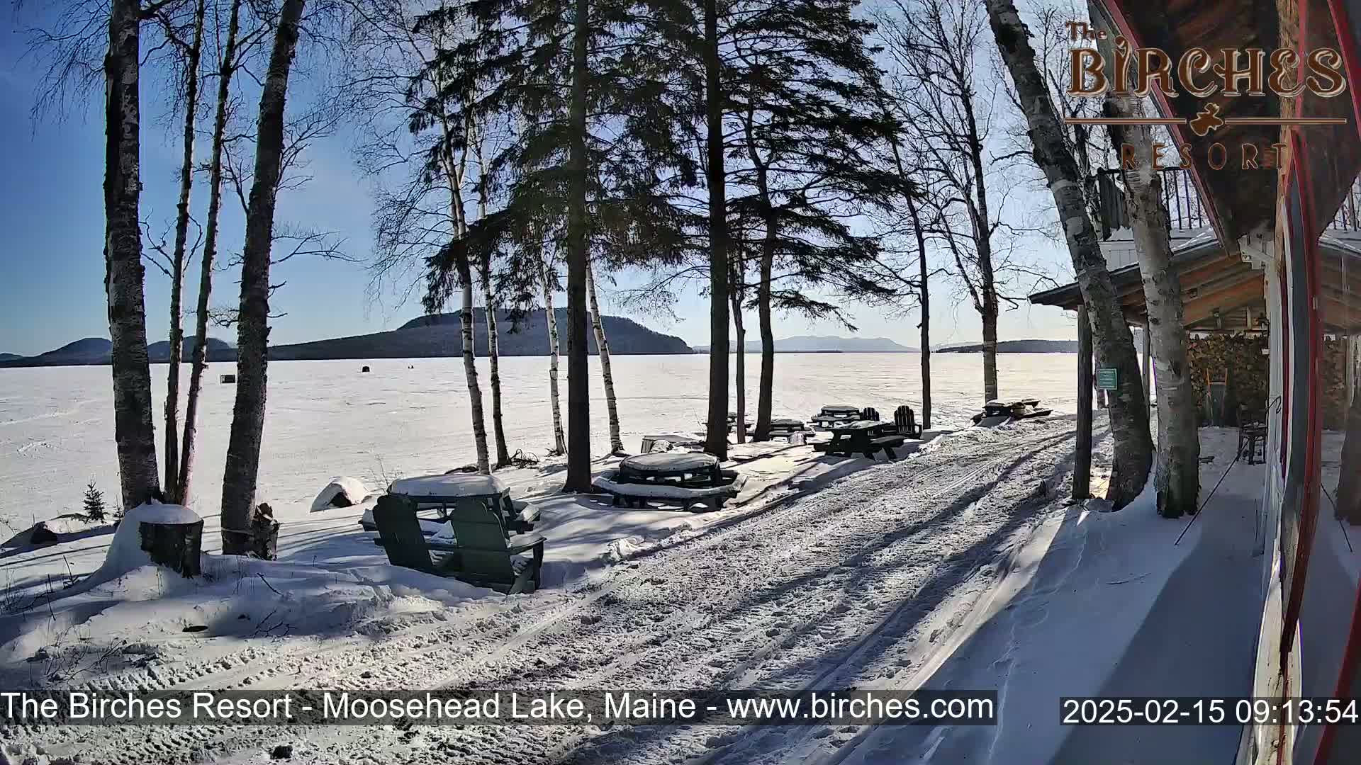 A snow-covered lakeside scene on a sunny day shows picnic tables and Adirondack chairs near a building, with a frozen lake and distant mountains in the background.