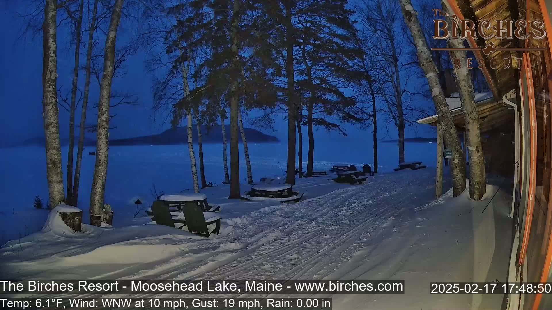 A snow-covered lakeside area at twilight with picnic tables and trees, under a dark blue sky with a calm lake in the background.