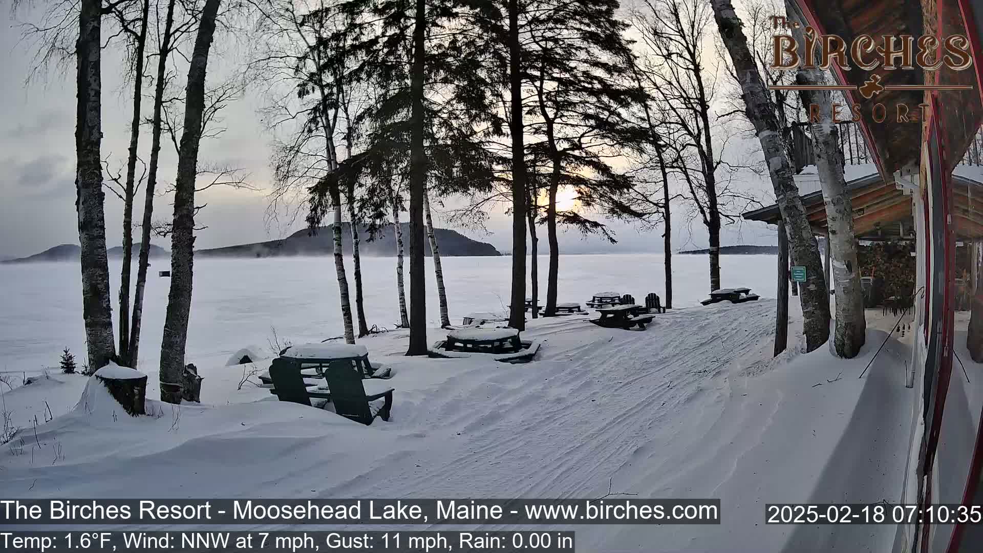 Snow-covered picnic tables sit near a lakeshore under a wintry sunrise, with a light fog over the frozen lake and bare trees in the foreground.