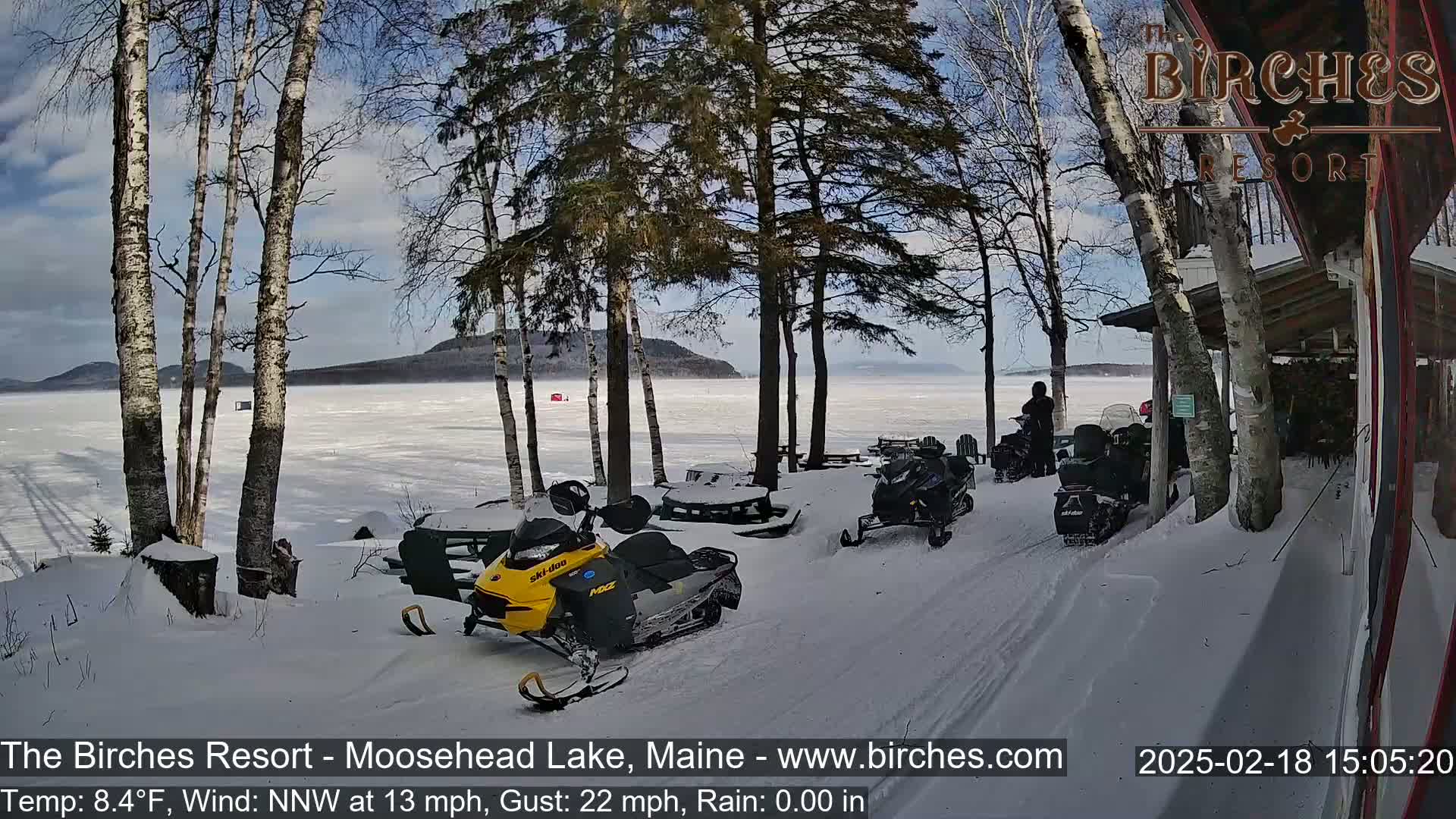 Several snowmobiles are parked near a snow-covered lakeshore with birch trees under a partly sunny sky, a moderate northwest wind, and a temperature of 8.4°F.