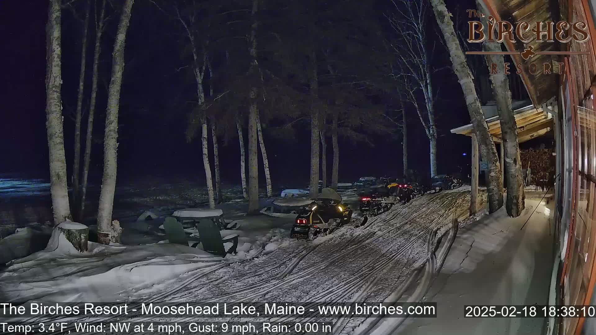 A snow-covered area at night, illuminated by artificial lights, shows several snowmobiles parked near a building with birch trees in the background, under a dark sky with a northwest wind.