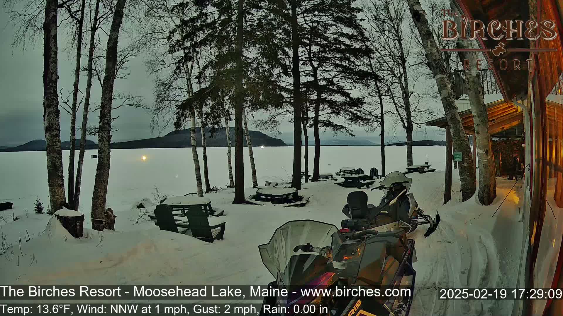 A snow-covered lakeside scene at dusk shows a snowmobile parked near picnic tables and trees, with a calm lake in the background.
