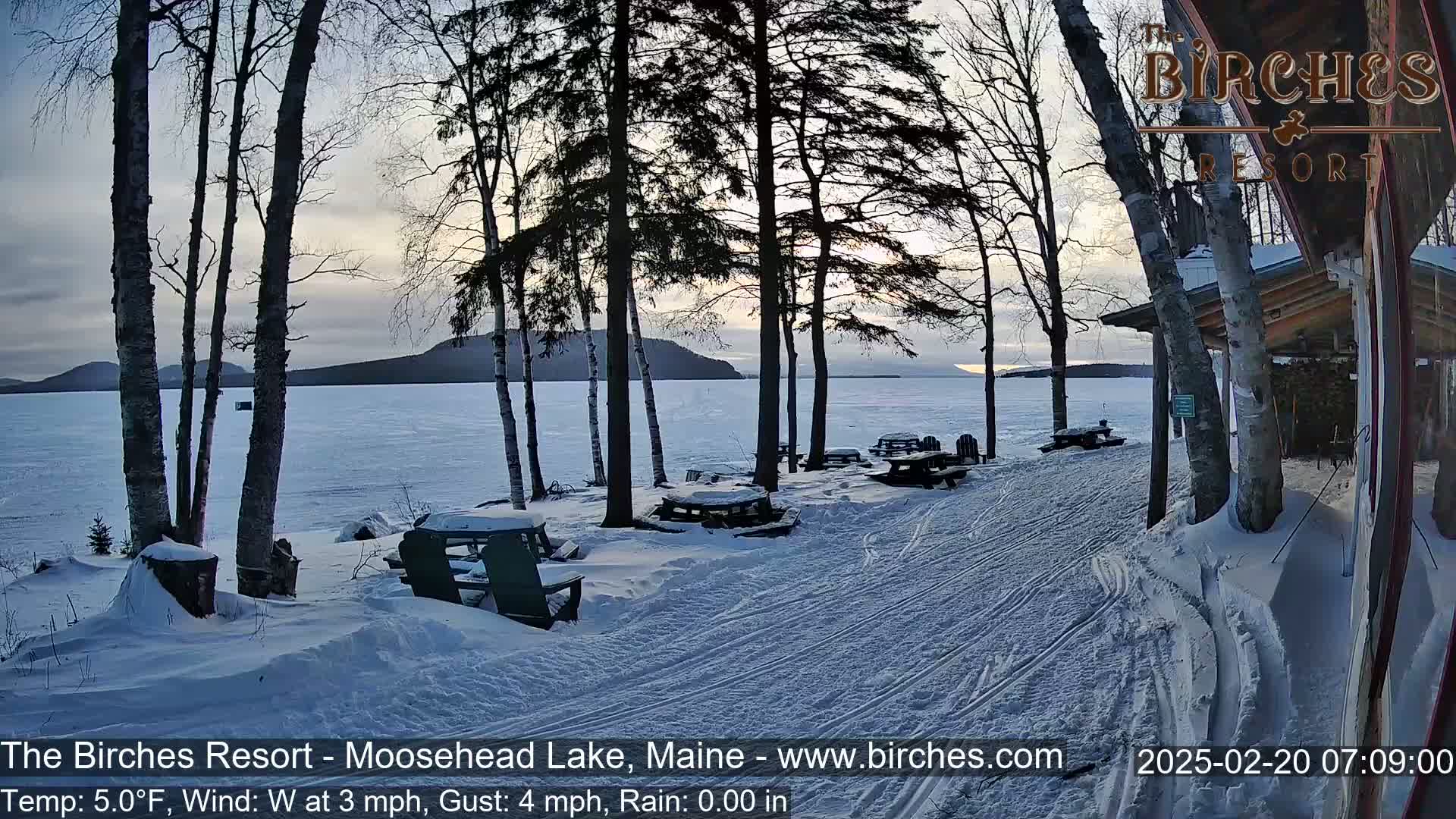 A snow-covered lakeside scene at sunrise shows picnic tables and chairs under bare trees near a building, with a calm frozen lake and distant hills in the background.