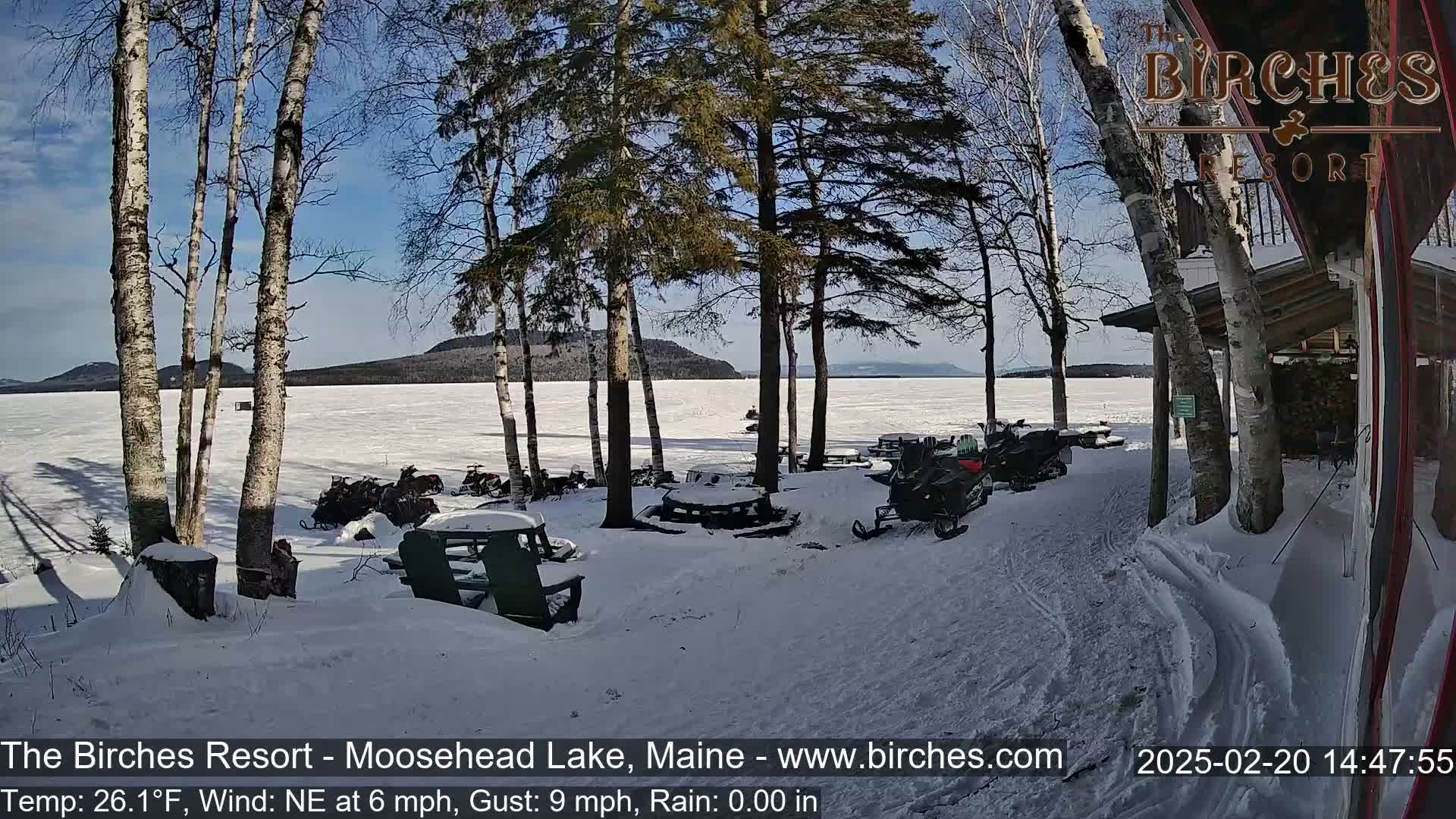 Several snowmobiles are parked near picnic tables in a snowy area beside a frozen lake under a sunny sky.