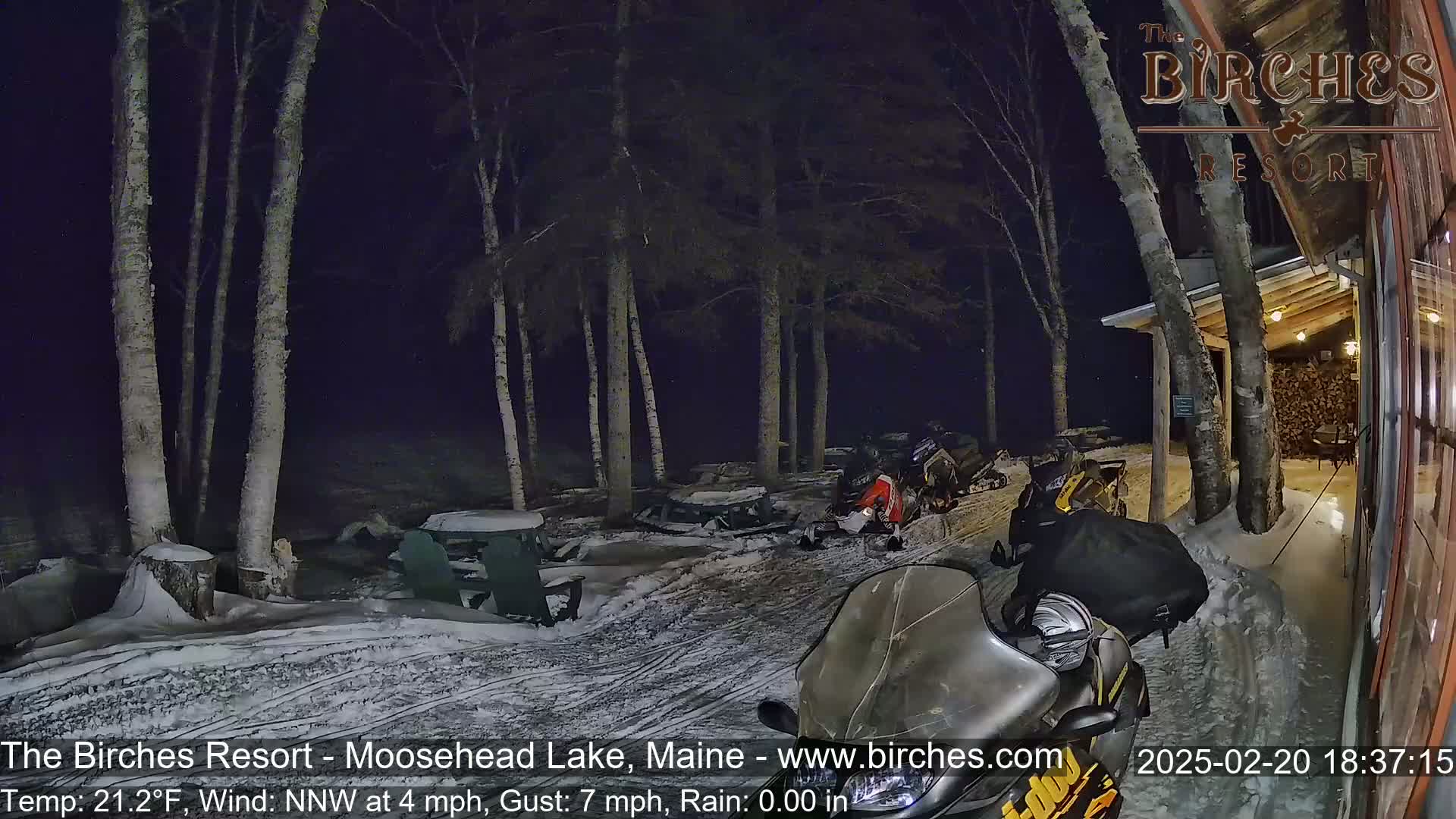 Several snowmobiles are parked in a snowy area next to a building under a dark, nighttime sky with light rain and a northwest wind.