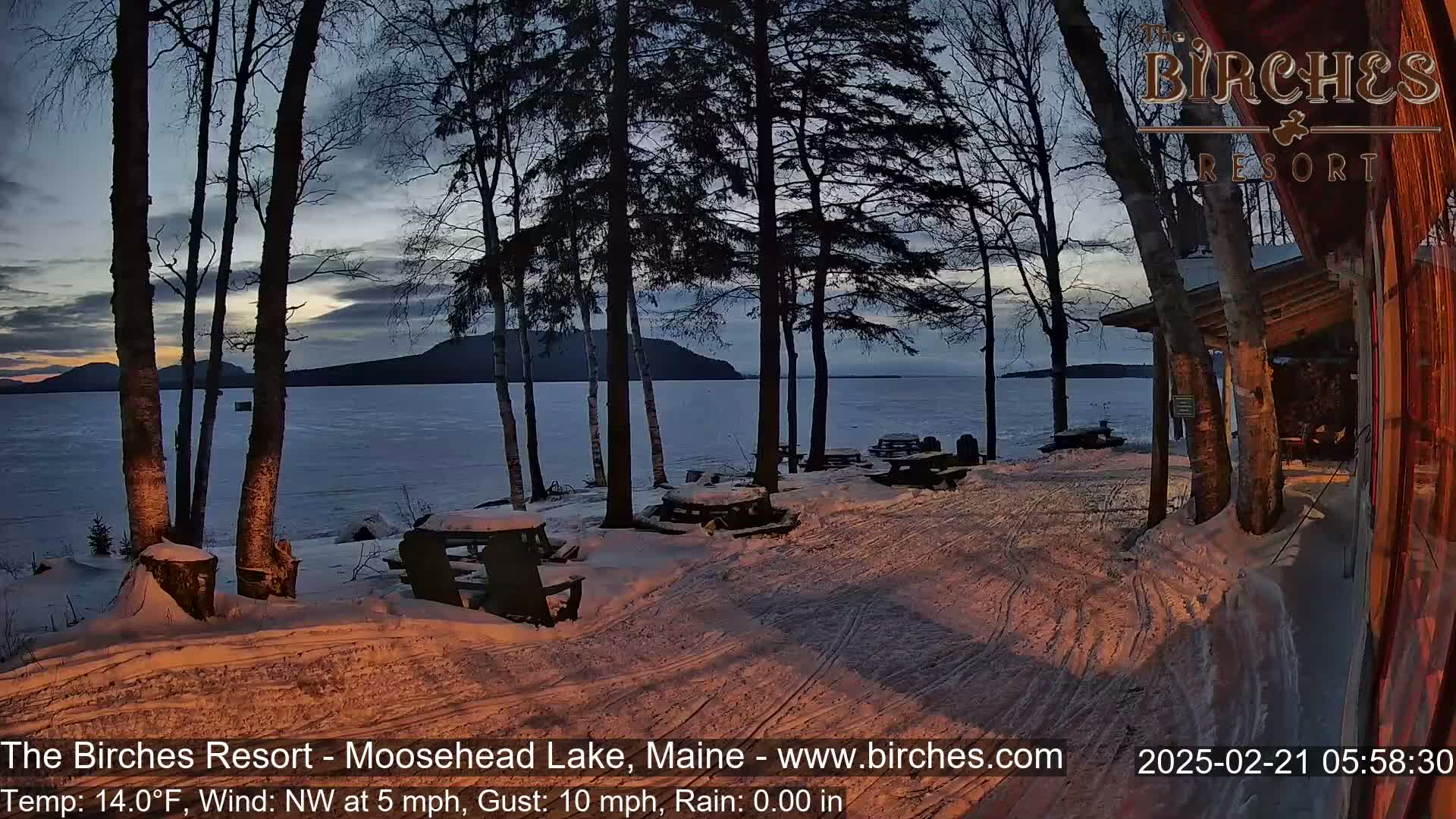 A snow-covered lakeside area with picnic tables and several birch trees at twilight, under a partly cloudy sky.