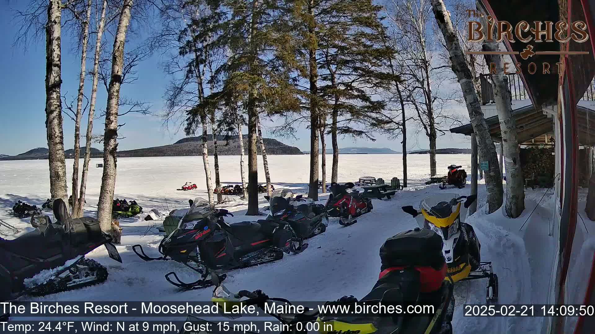 Several snowmobiles are parked near a snow-covered lake shore under a sunny sky, with birch trees and a wooden building visible.