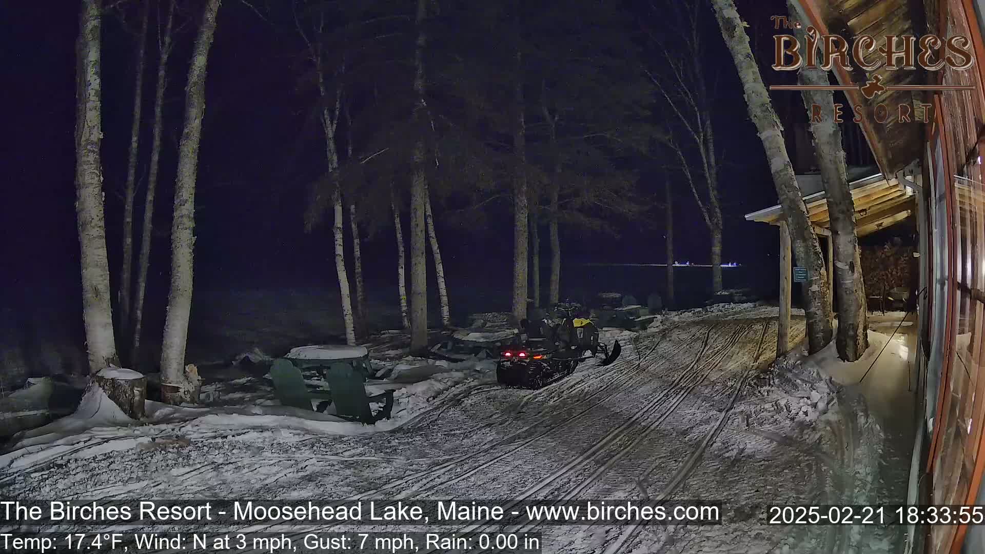 A snowmobile sits parked in a snow-covered area next to a building at night, under a dark sky with a calm wind.