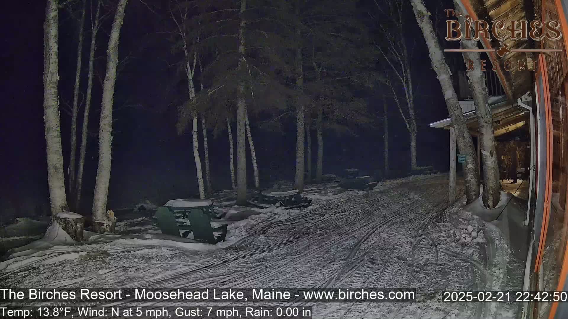 A snow-covered area with picnic tables and chairs near a building at night, under a dark sky with a north wind.