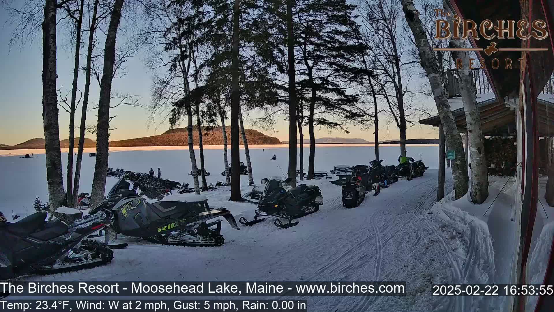 Several snowmobiles are parked near a lake shore, surrounded by trees in a snowy landscape at sunset.