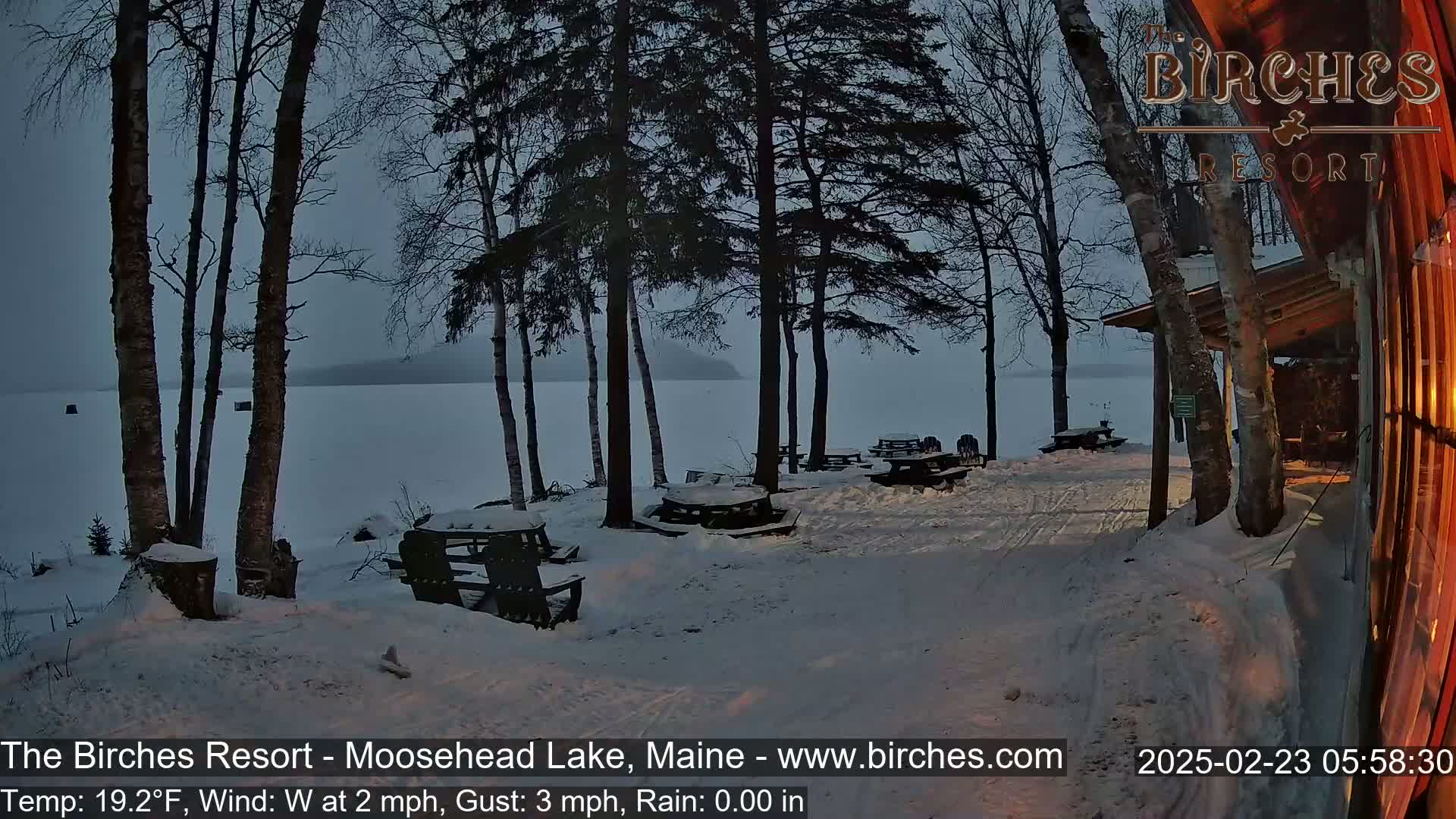 A snow-covered lakeside area with several empty picnic tables, trees, and a building is visible under a dusky sky.