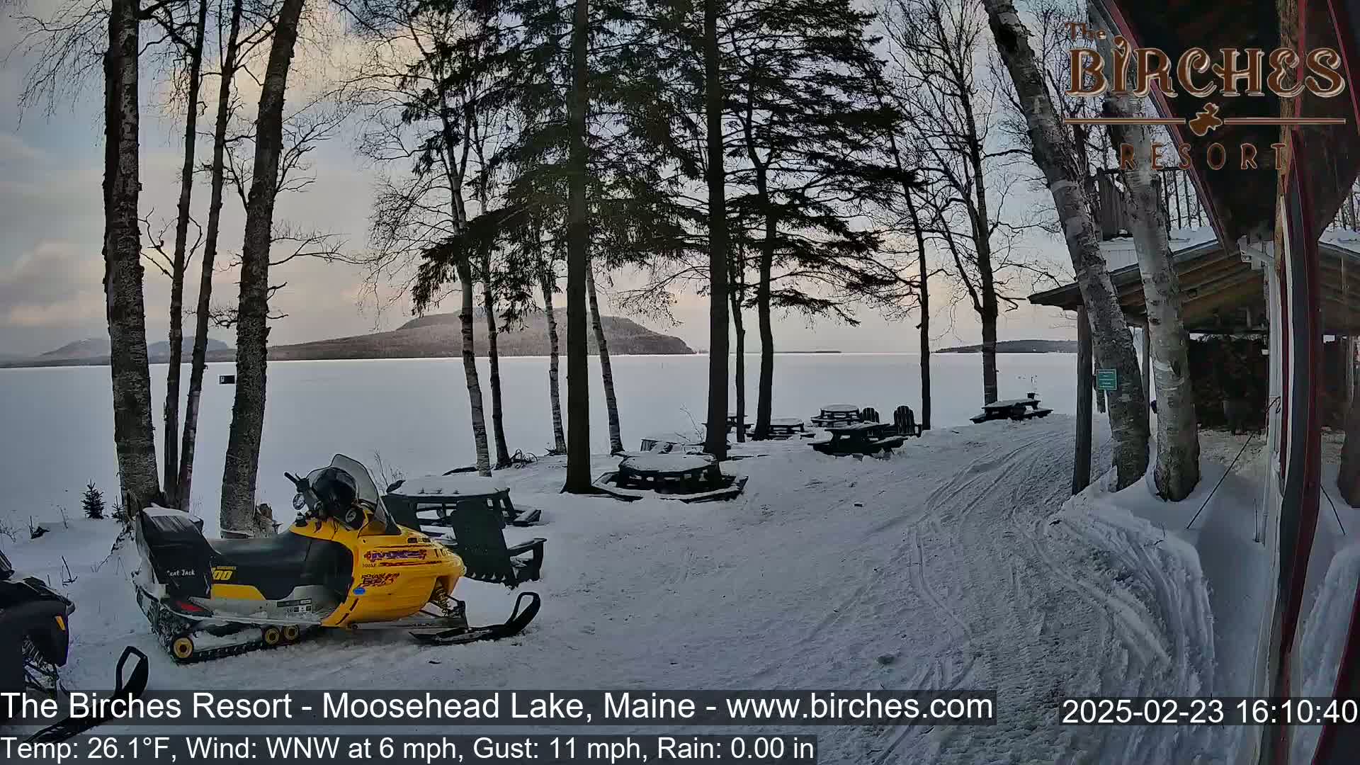 A snow-covered lakeside area with picnic tables, a snowmobile, and trees, under a partly cloudy sky.