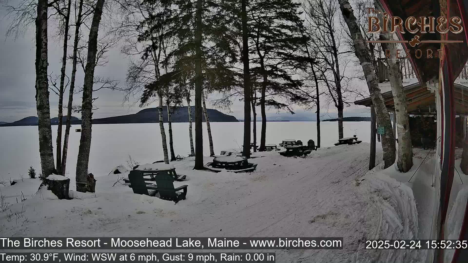 A snow-covered lakeside picnic area with several empty tables and chairs is visible near a building, overlooking a frozen lake under a cloudy sky.