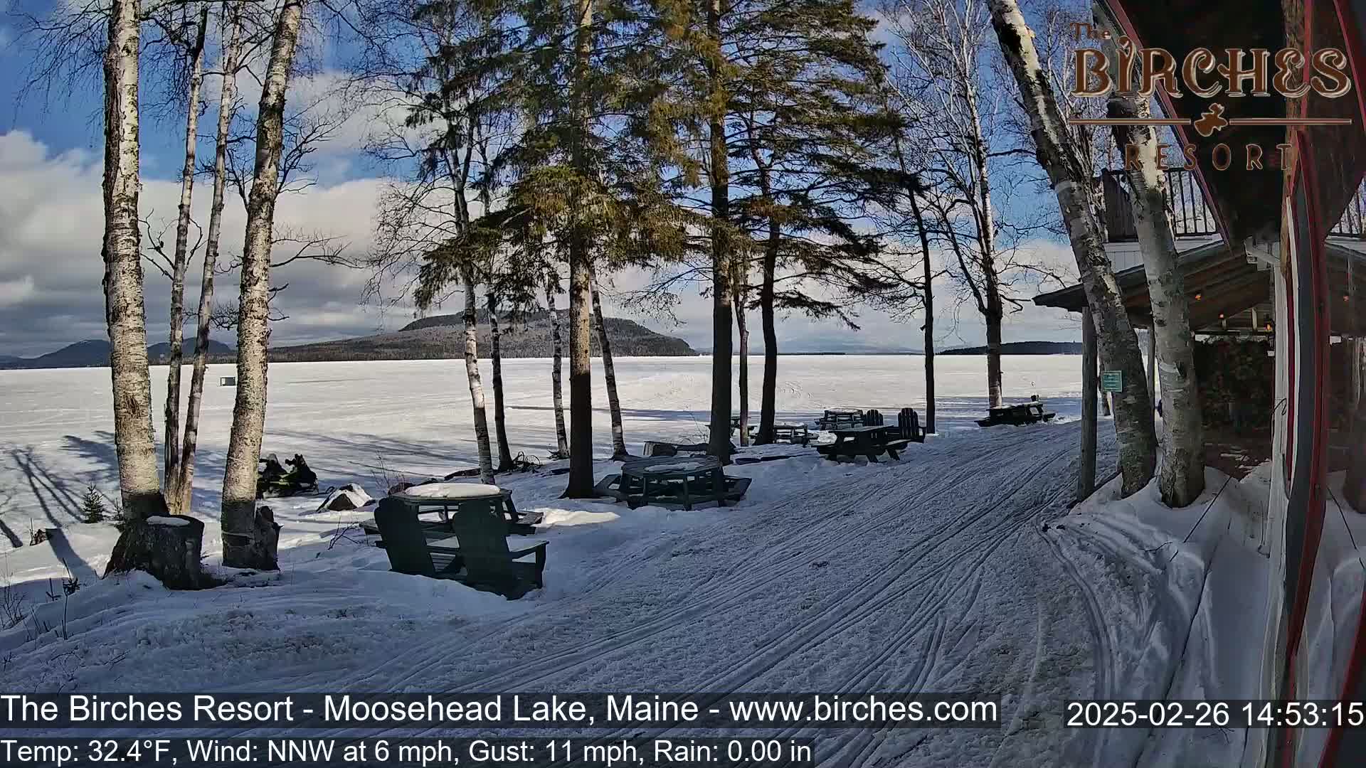 A snowy lakeside scene with picnic tables and snowmobiles under a partly sunny sky.