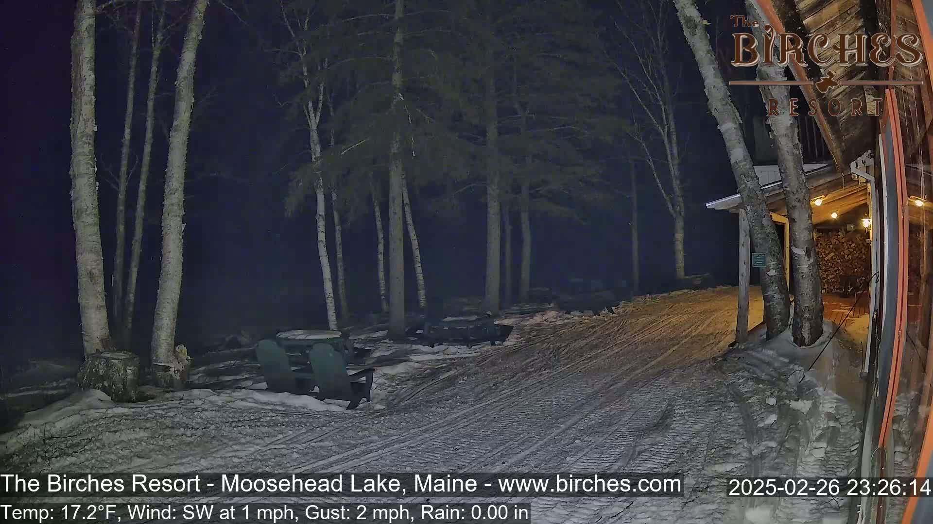 A snow-covered outdoor area at night, featuring several birch trees, picnic tables, and Adirondack chairs near a building, under a southwest wind of 1 mph with gusts up to 2 mph and no rain.