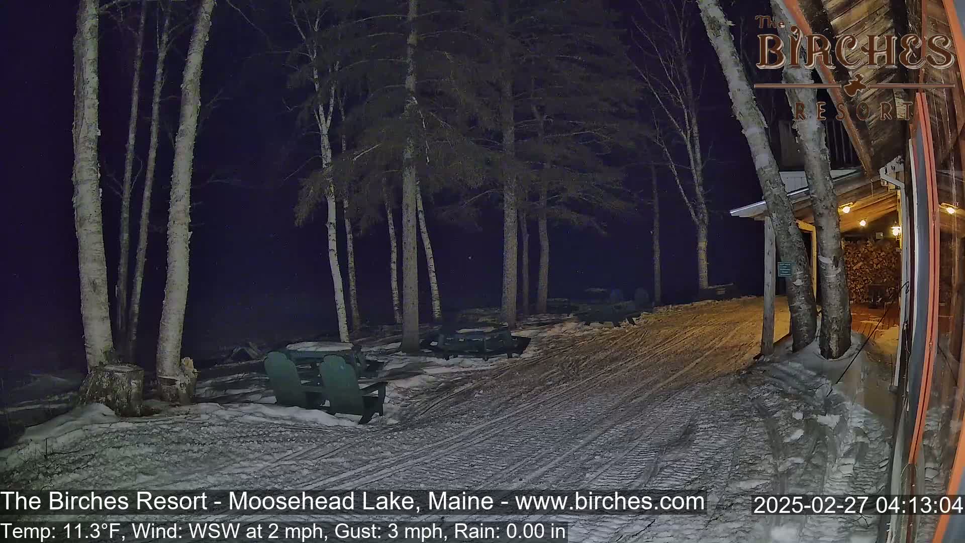 A snowy, nighttime scene shows a resort patio area with several picnic tables and chairs under birch trees, lightly raining with a 2 mph wind.