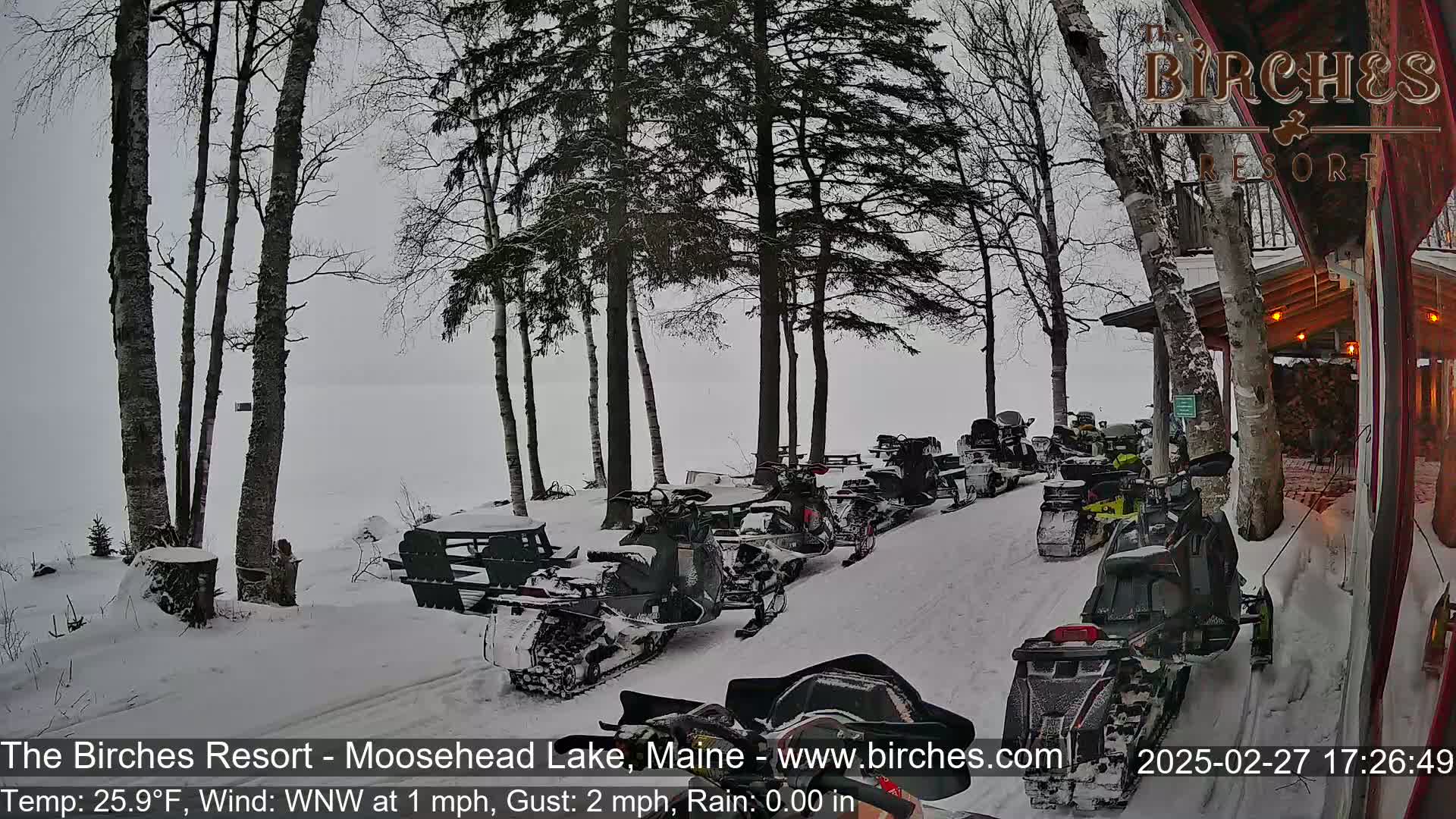 A snowy scene shows a line of snowmobiles parked near a lakeside building under a gray sky with light rain.