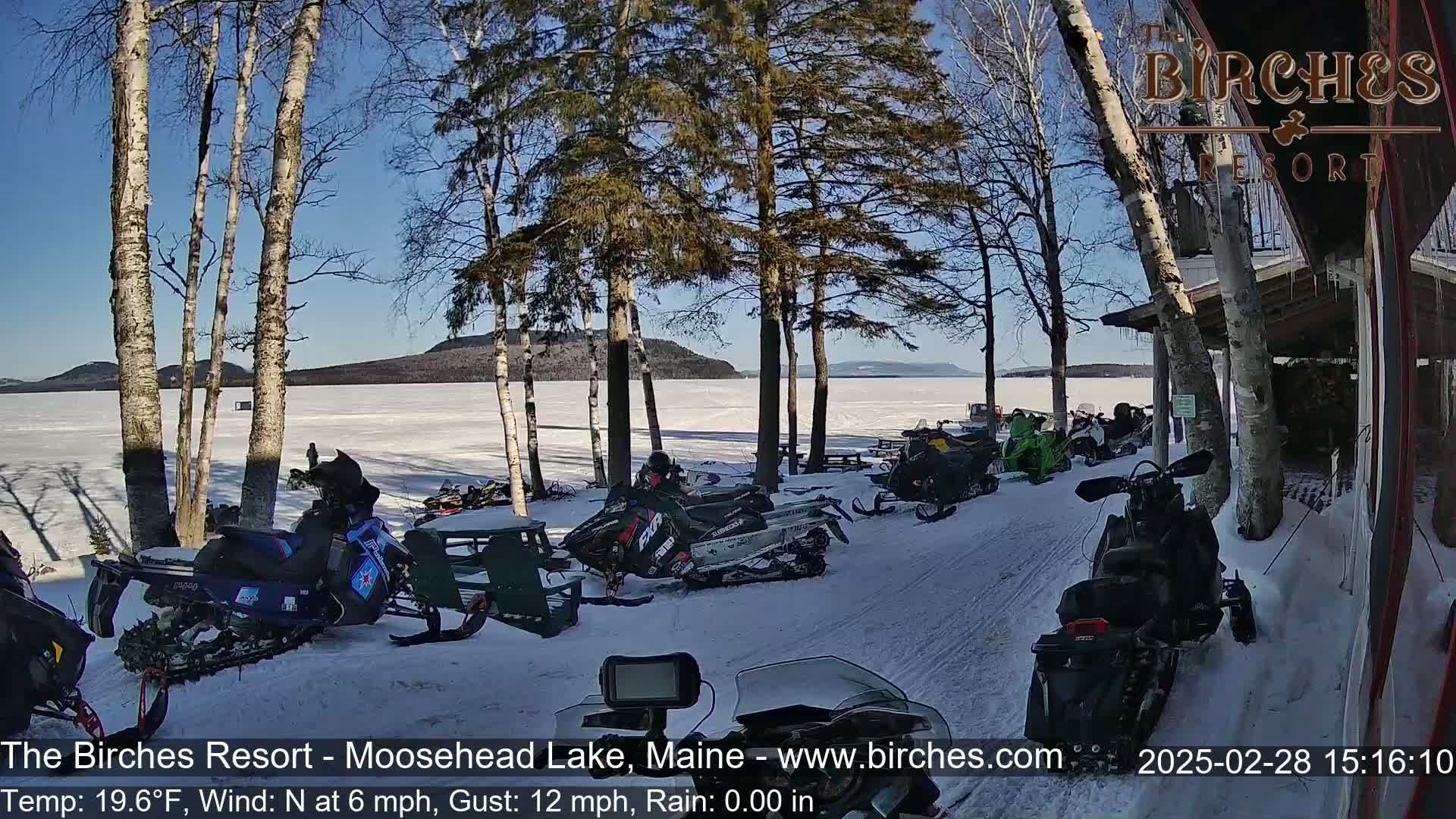 Several snowmobiles are parked near a lakeside building on a sunny, snowy day.