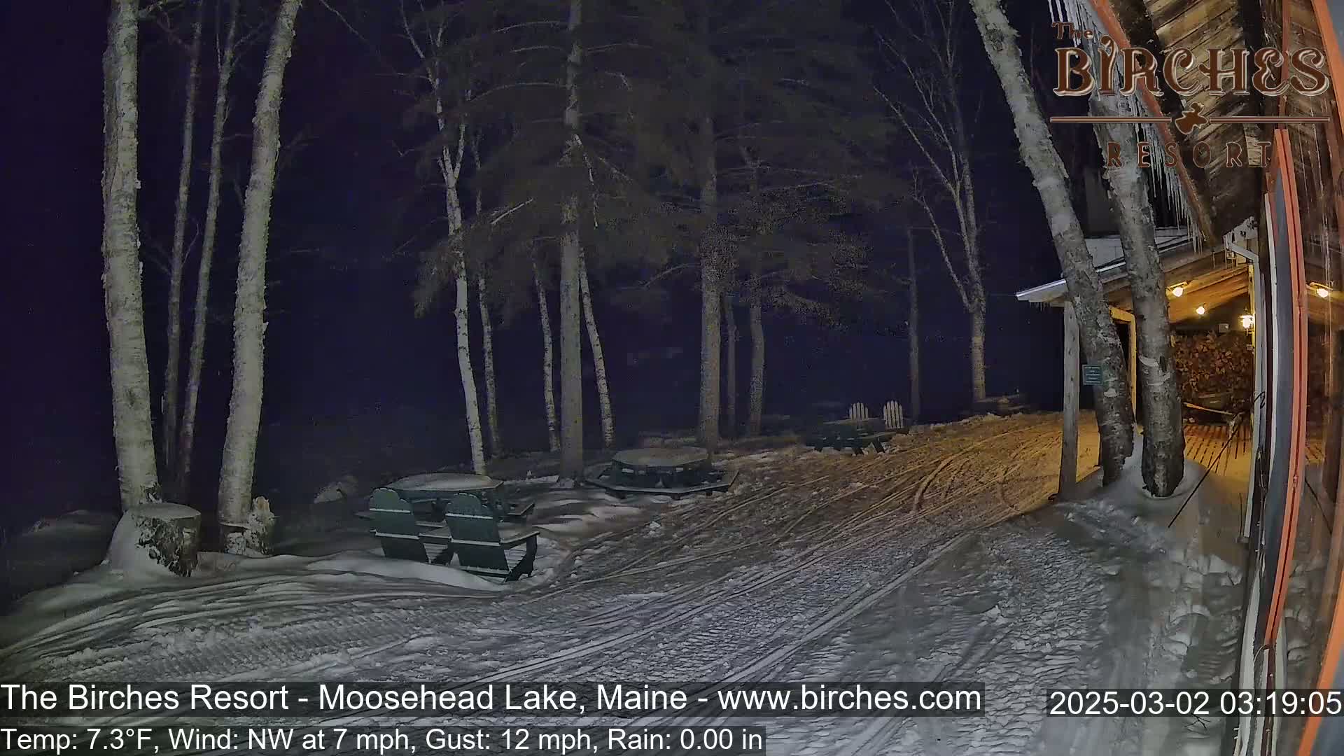 A snow-covered outdoor area at night, illuminated by lights from a nearby building, shows several picnic tables and chairs under tall birch trees in a 7.3°F temperature with a northwest wind.