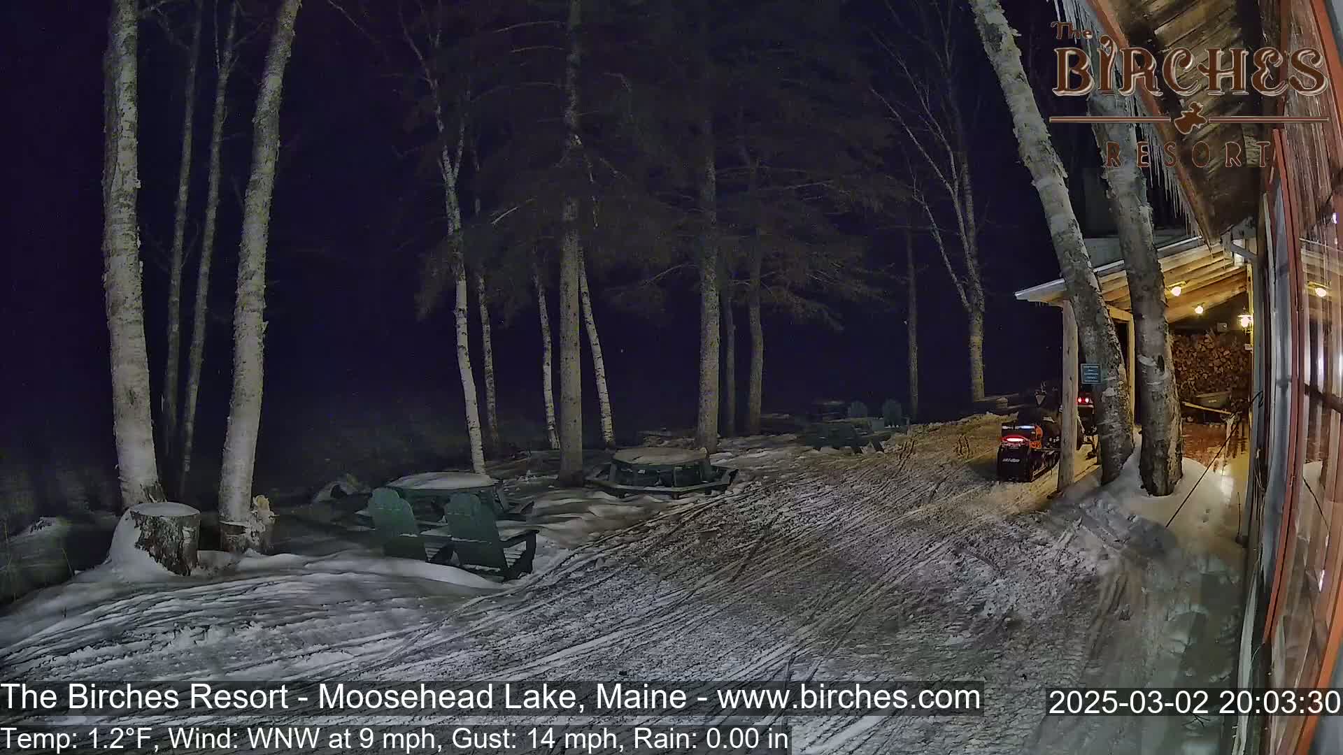 A snow-covered outdoor area at night, illuminated by lights, features picnic tables and chairs under birch trees, with a snowmobile parked near a building.