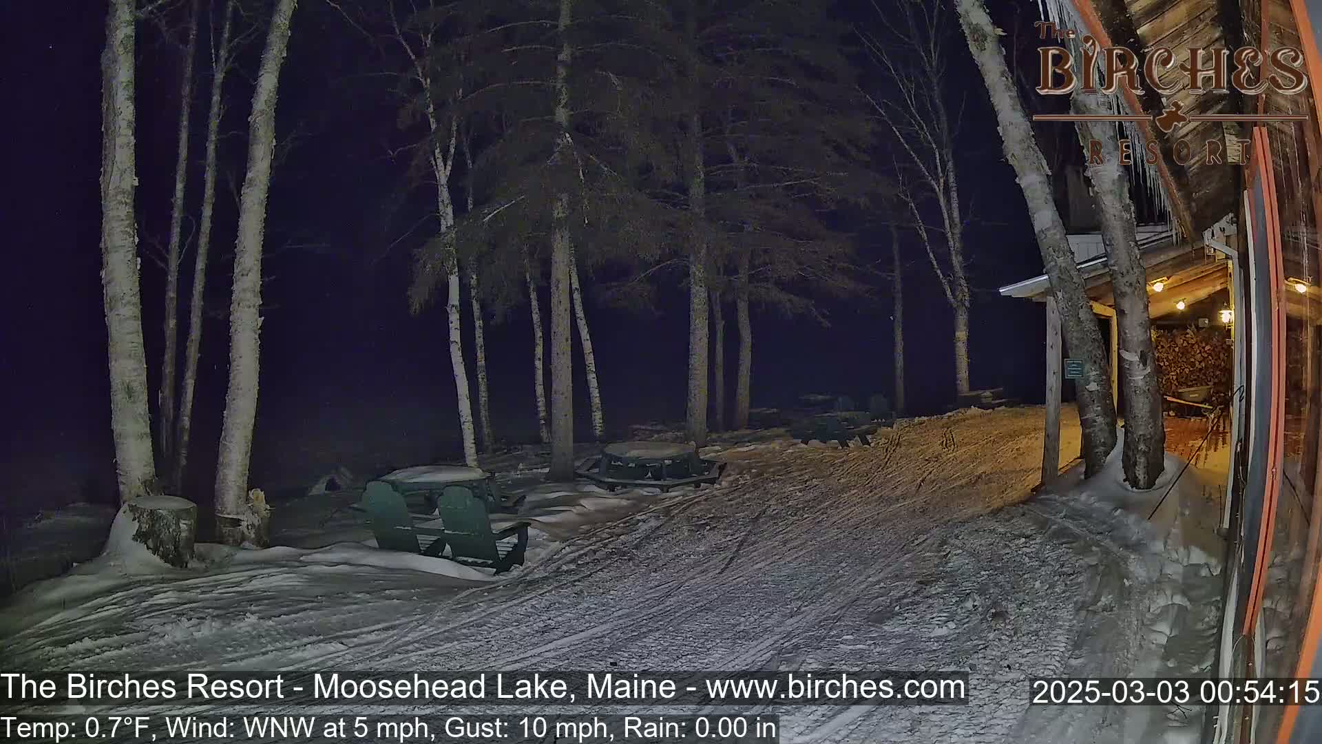 A snow-covered outdoor area at night, illuminated by lights from a building, shows picnic tables and chairs under several birch trees in a 5 mph wind with a 0.7°F temperature.