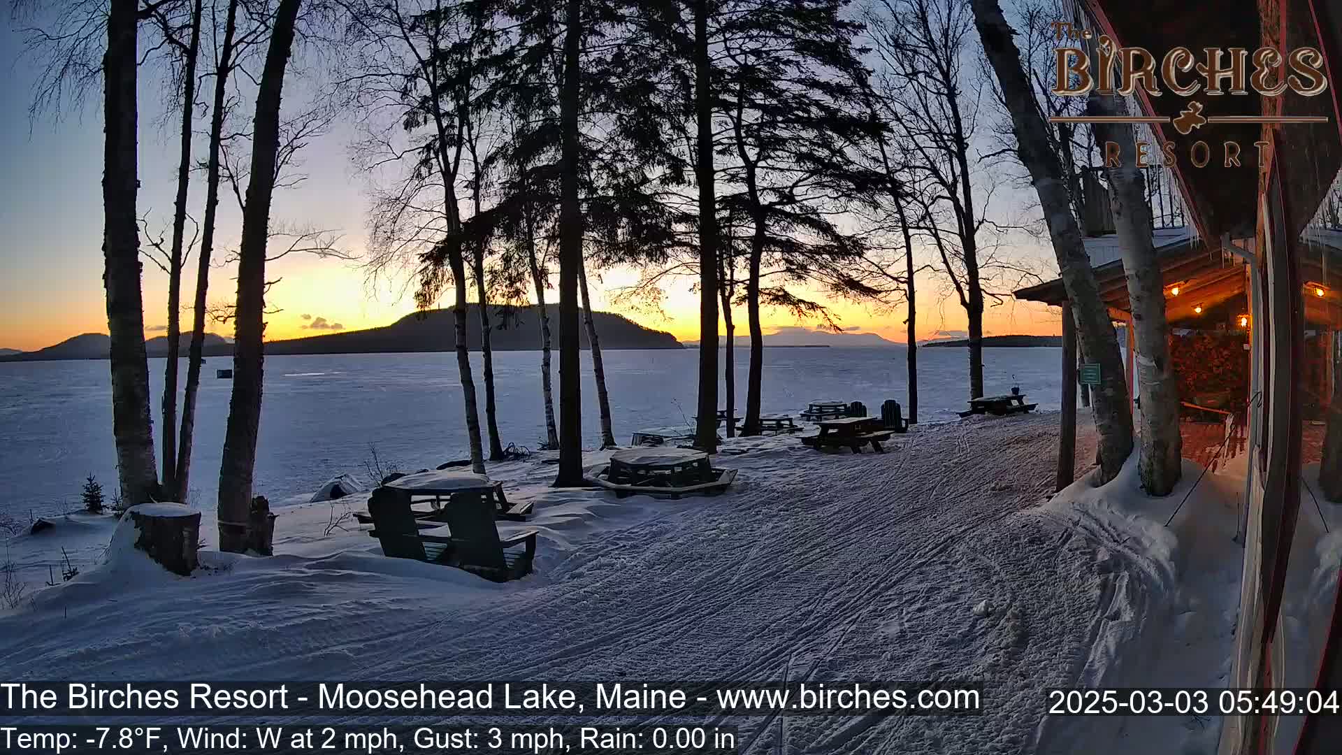 A snow-covered lakeside patio with picnic tables and chairs at sunset, overlooking a frozen lake and distant mountains.