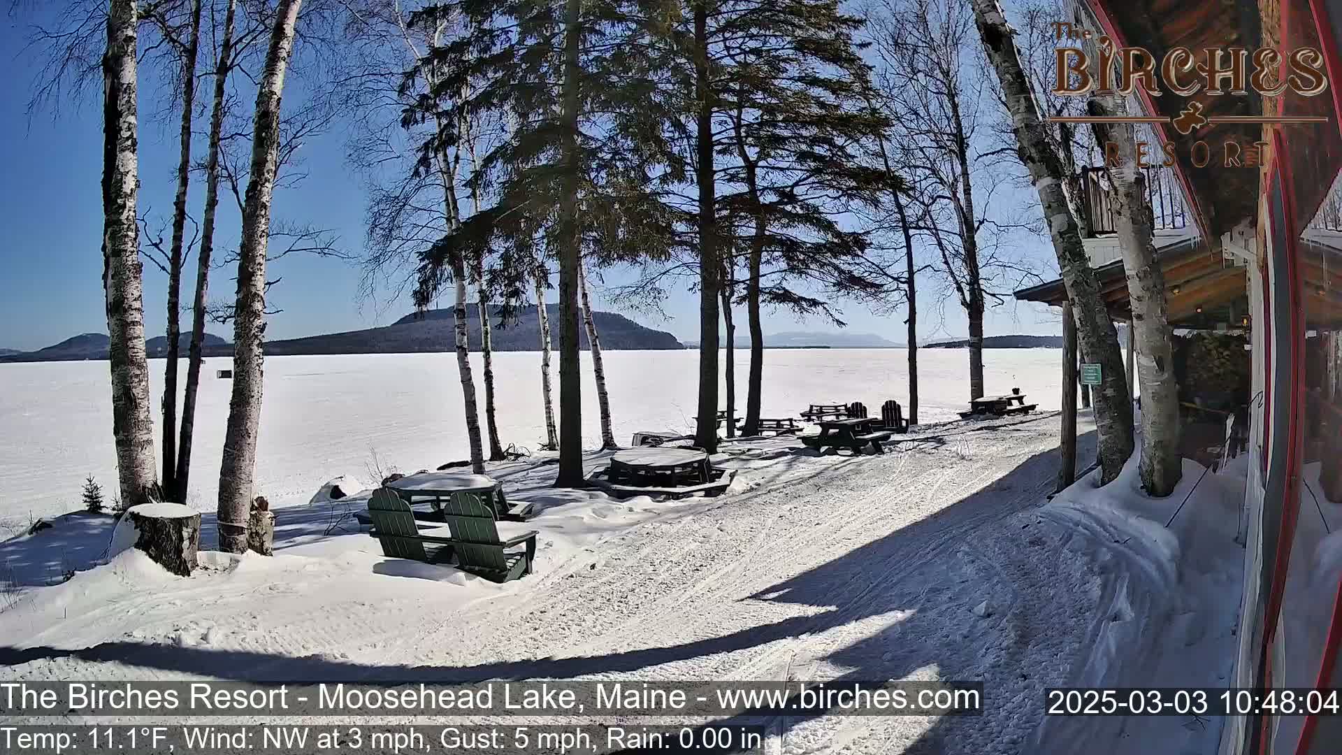 A snow-covered lakeside patio with picnic tables and Adirondack chairs, under a sunny sky, overlooks a frozen lake and distant mountains.
