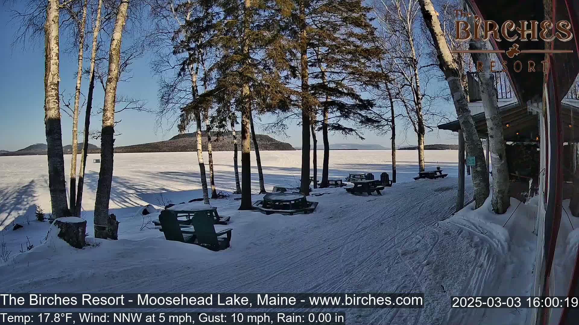 A snow-covered lakeside patio with picnic tables and chairs under a sunny sky, overlooking a frozen lake and distant hills.