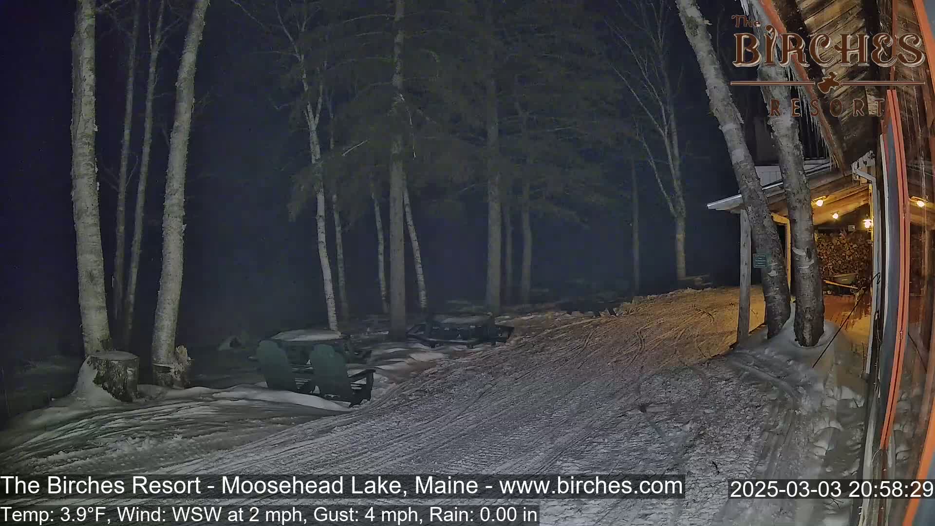A snow-covered area with birch trees at night, illuminated by dim light, shows a partially visible building and some outdoor furniture.