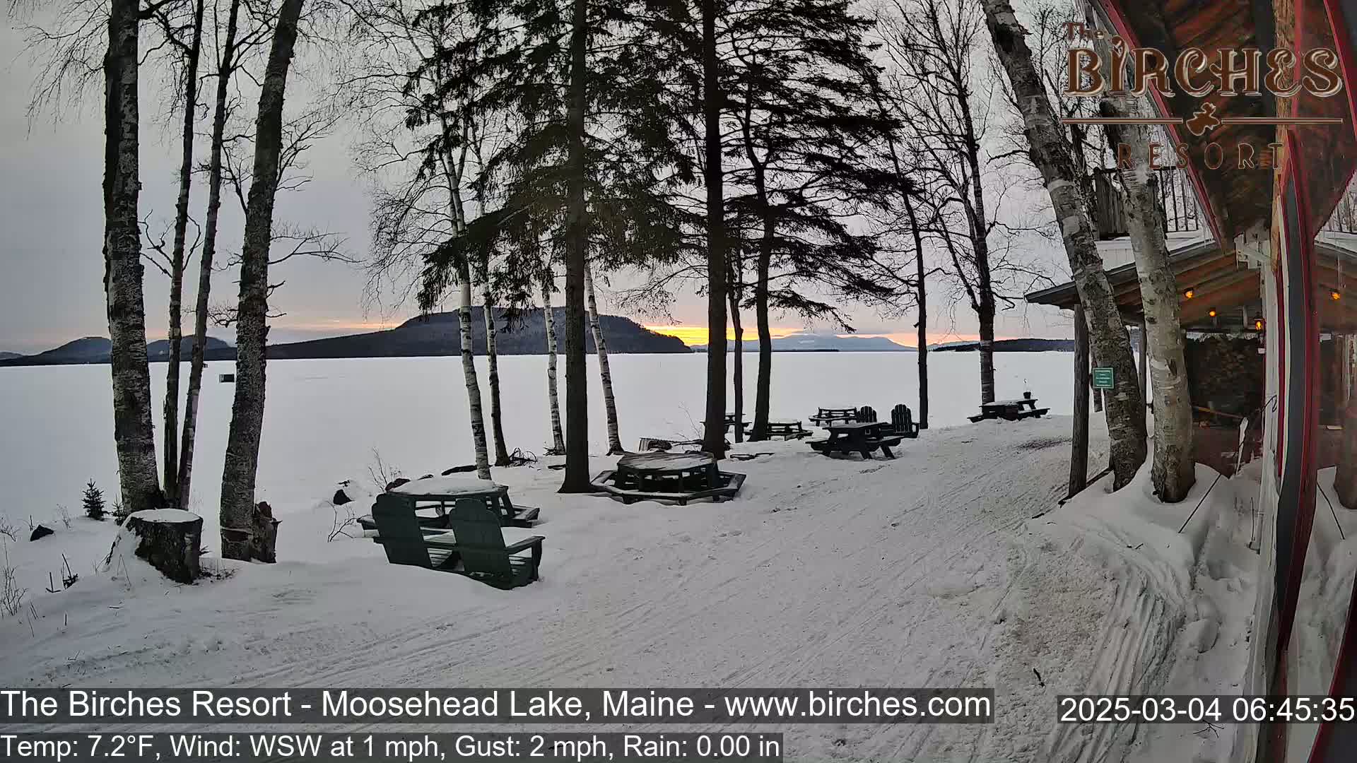A snow-covered lakeside area with picnic tables and trees at sunset, under a mostly clear sky.