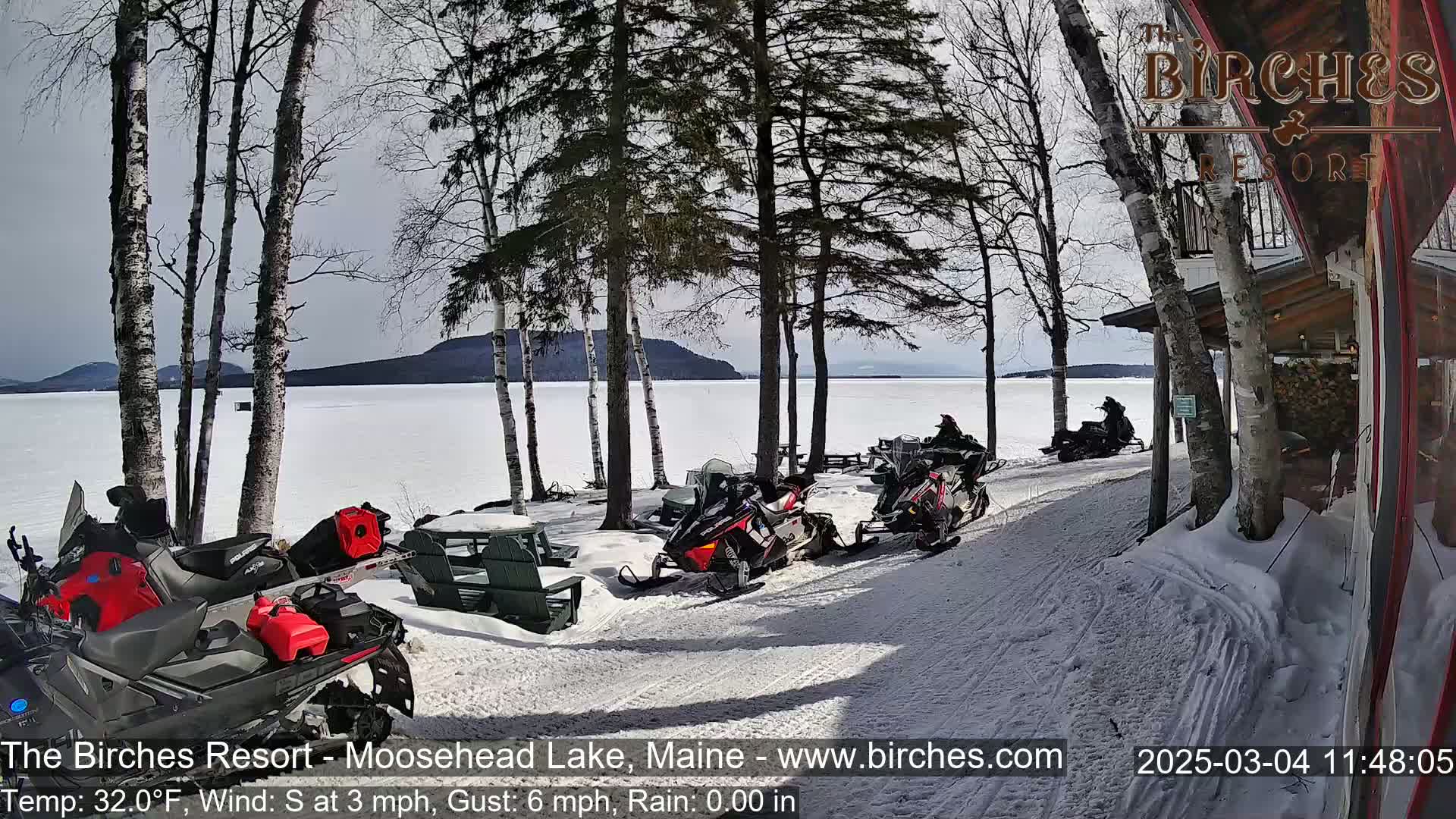 Several snowmobiles are parked near a lakeside lodge on a sunny, snowy day with a light south wind.
