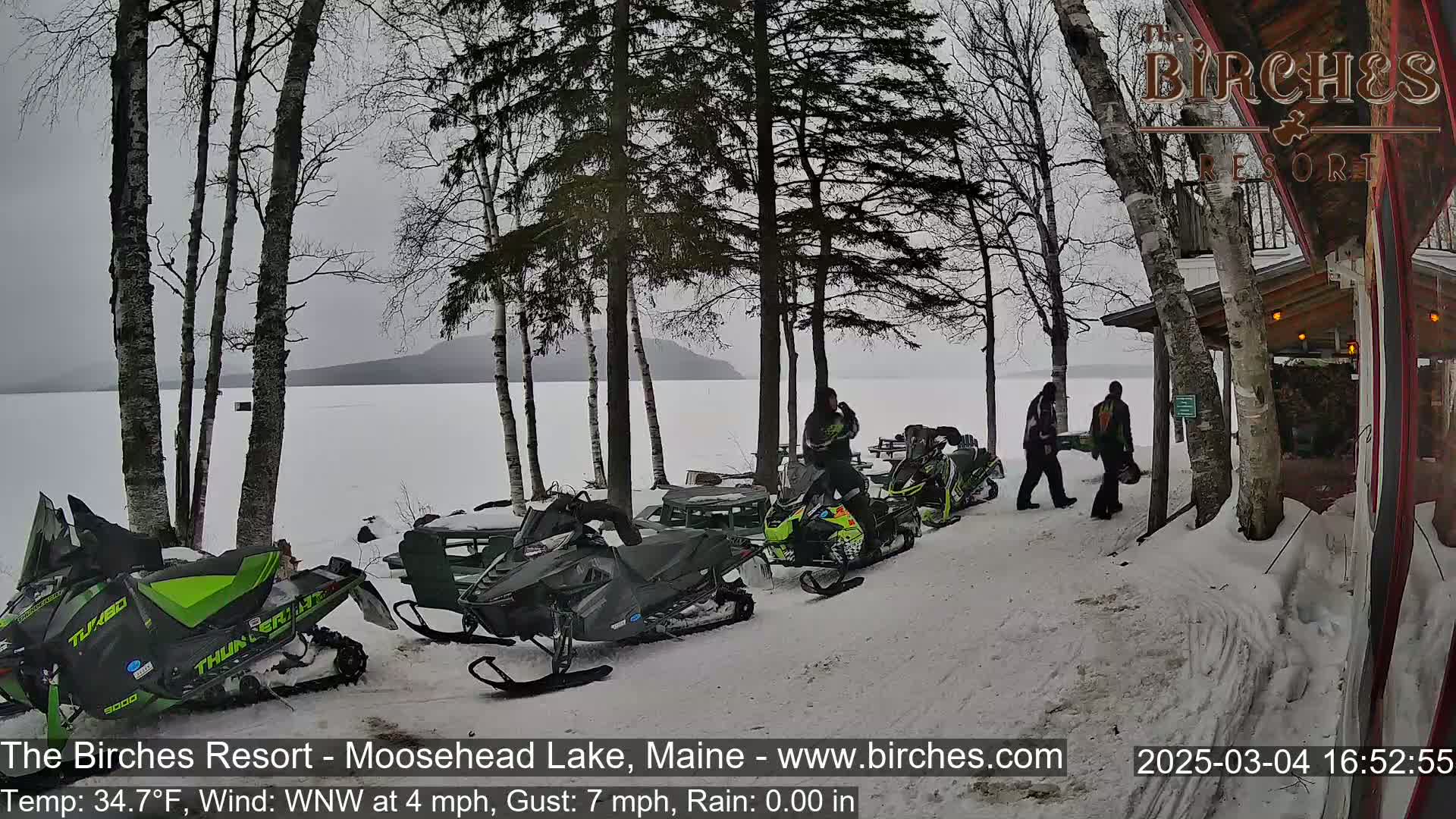 Several snowmobiles are parked near a lakeside lodge in snowy conditions with a light rain and overcast skies.
