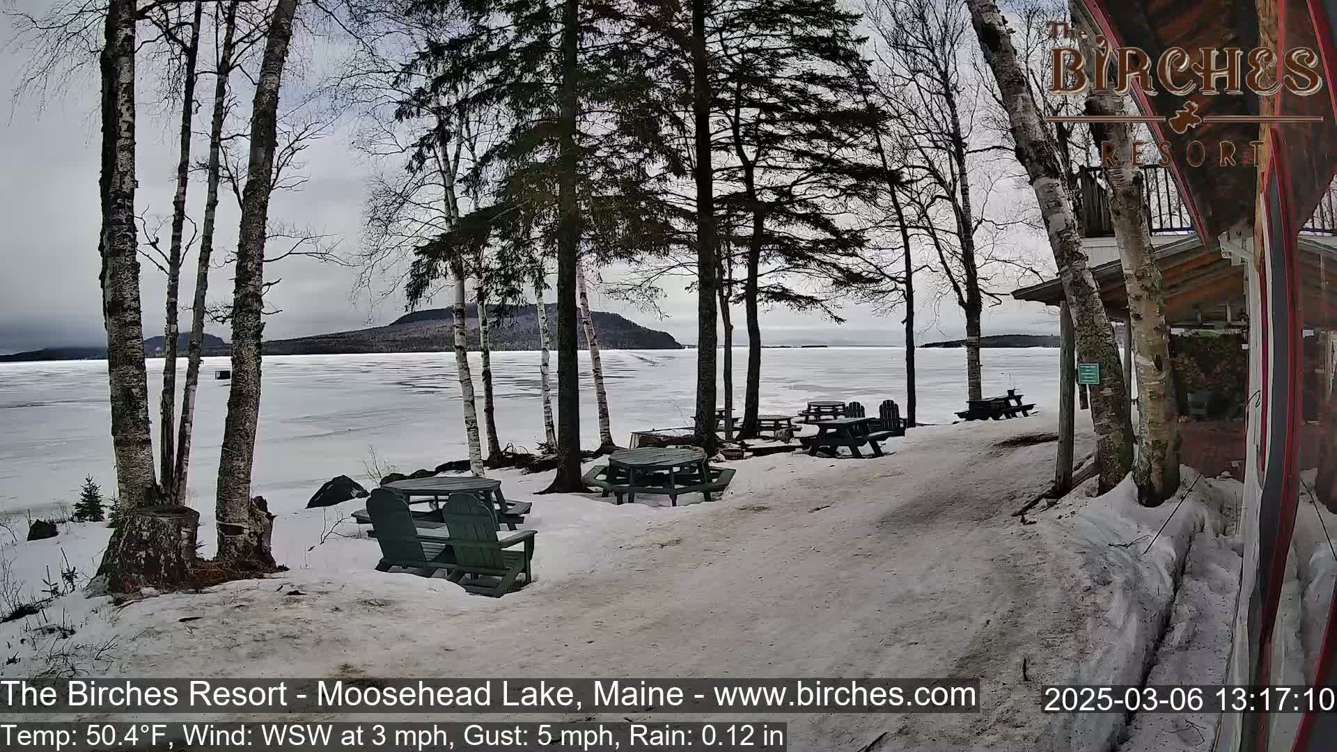 A snow-covered lakeside patio with picnic tables and chairs sits under a gray, overcast sky overlooking a partially frozen lake.