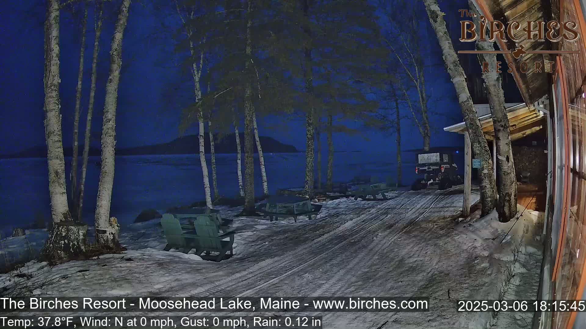 A snow-covered lakeside area at night, under a dark blue sky, shows empty picnic tables and Adirondack chairs near a building with birch trees in the foreground.