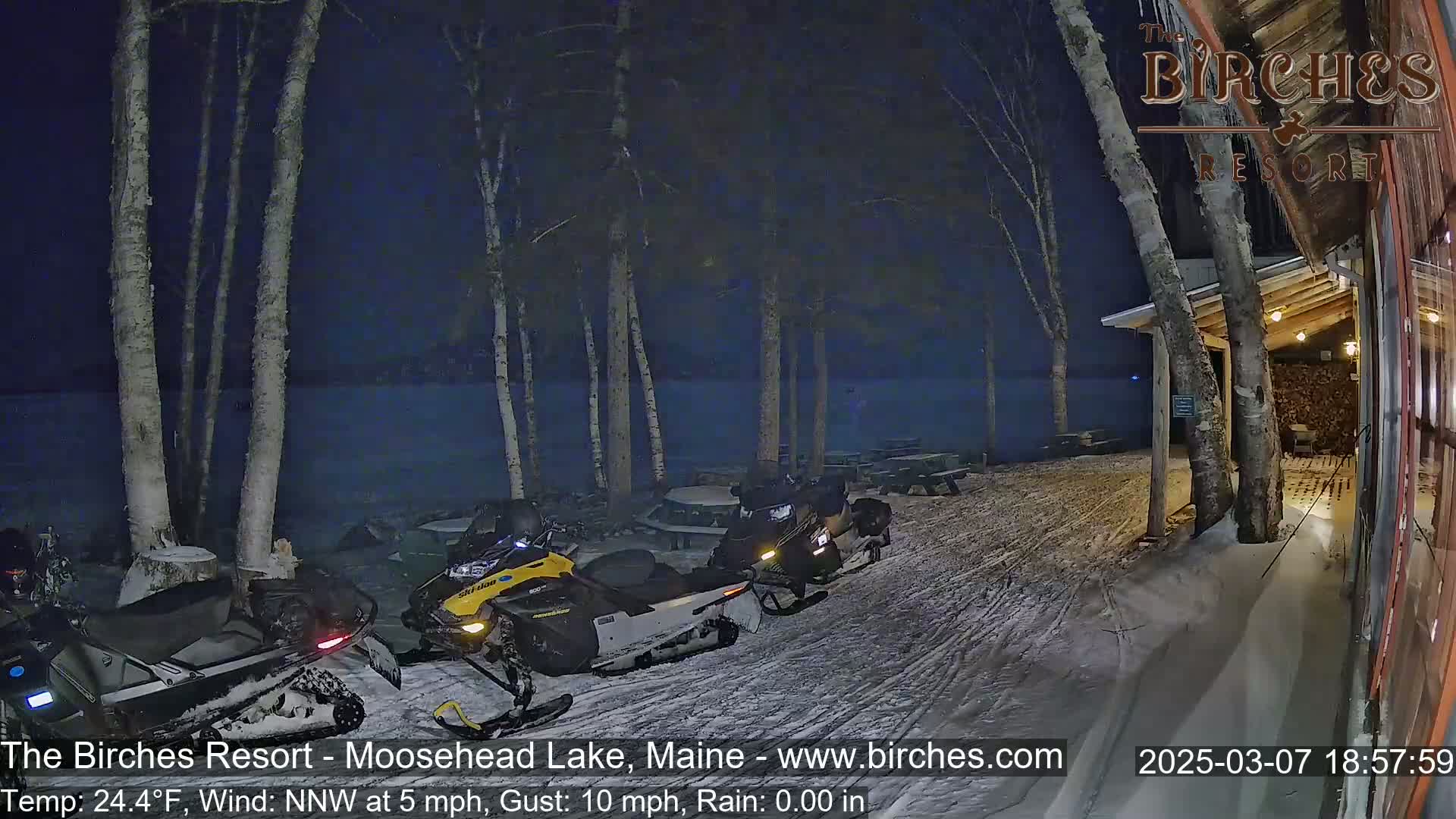 Several snowmobiles are parked outside a lakeside building at night in snowy conditions with a northwest wind.