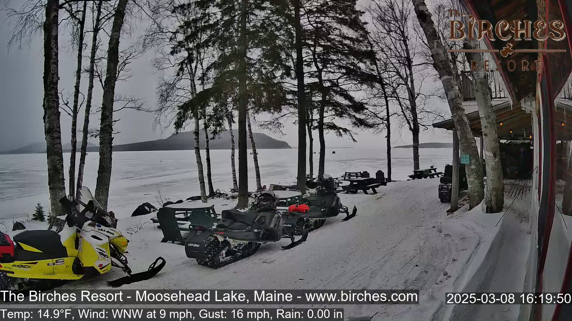 Several snowmobiles are parked near a lakeside resort on a snowy, overcast day with a wind of 9 mph and gusts up to 16 mph.