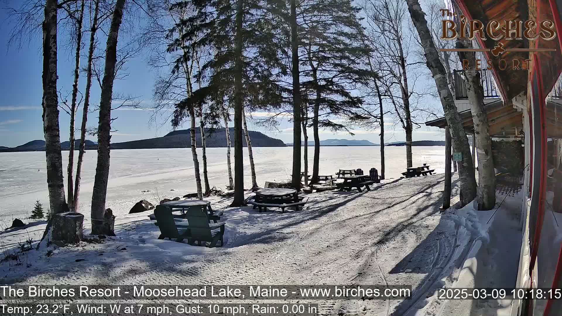 A sunny, snowy lakeside scene shows several picnic tables and chairs near a building, with a frozen lake and distant mountains in the background.