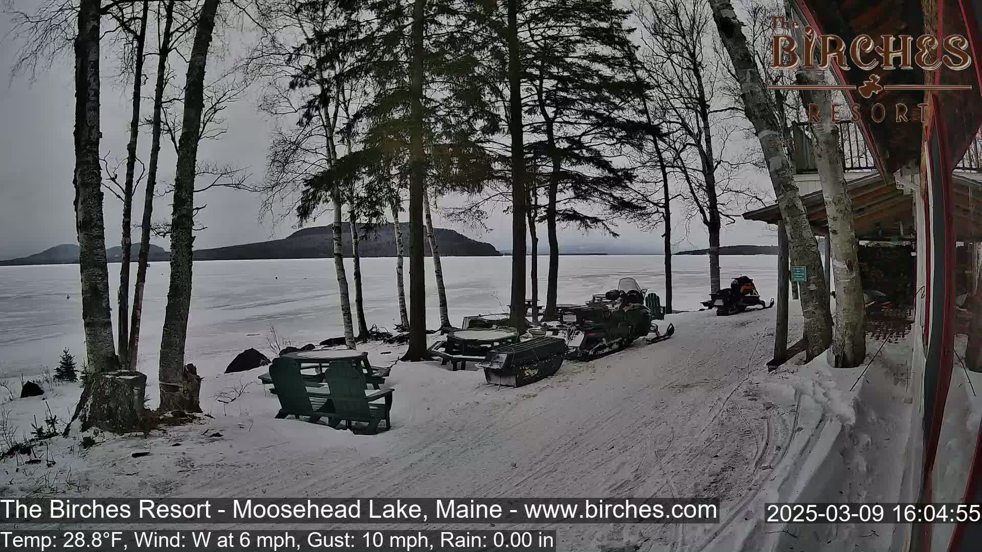 Snow-covered ground near a frozen lake, with several snowmobiles parked near picnic tables under bare trees on an overcast day.