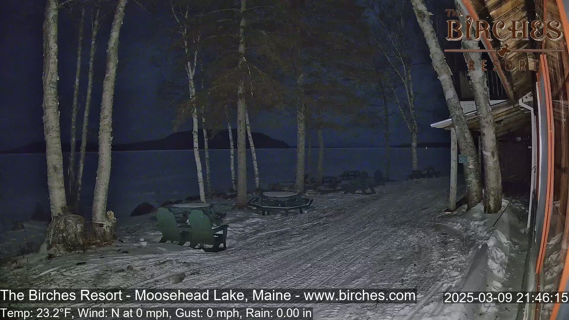 A snow-covered lakeside area at night shows picnic tables and chairs under birch trees near a building, with calm water visible in the background.