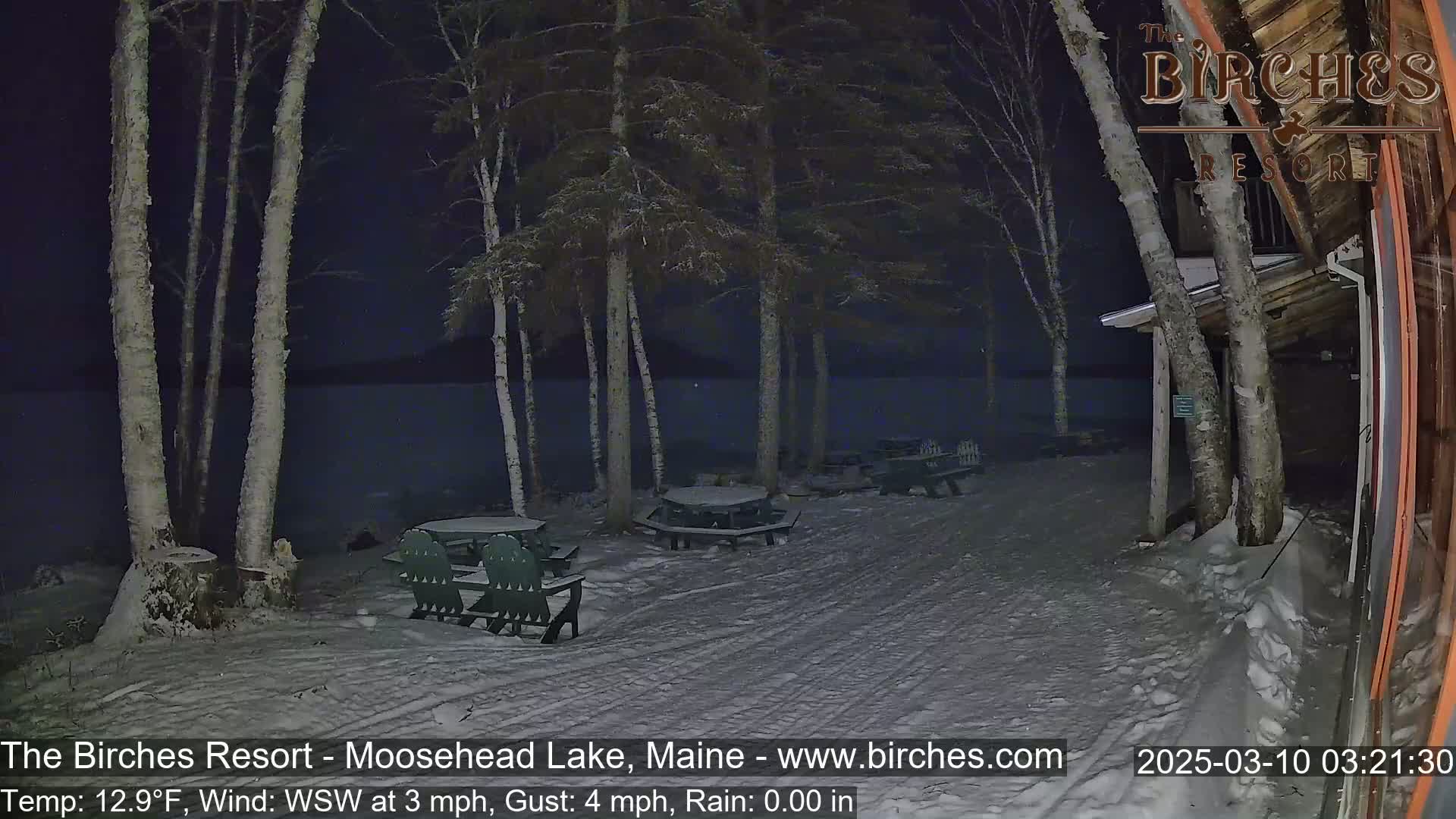 A snow-covered lakeside area at night, illuminated by a dim light, features several picnic tables and chairs near a building under a dark sky with a light wind.