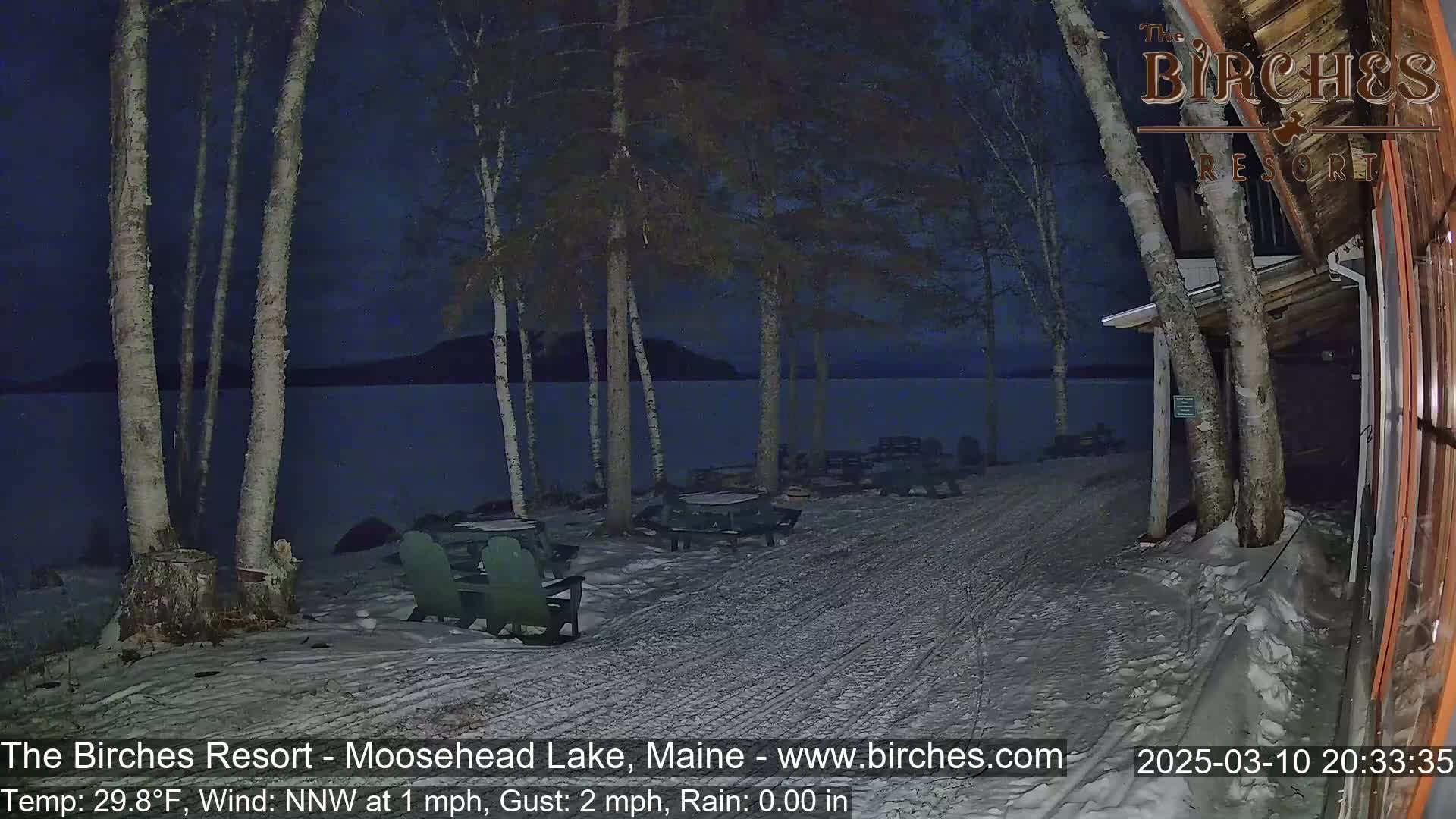 A snow-covered lakeside area at night, featuring several picnic tables and chairs near birch trees, with a calm lake visible in the background under a dark sky.