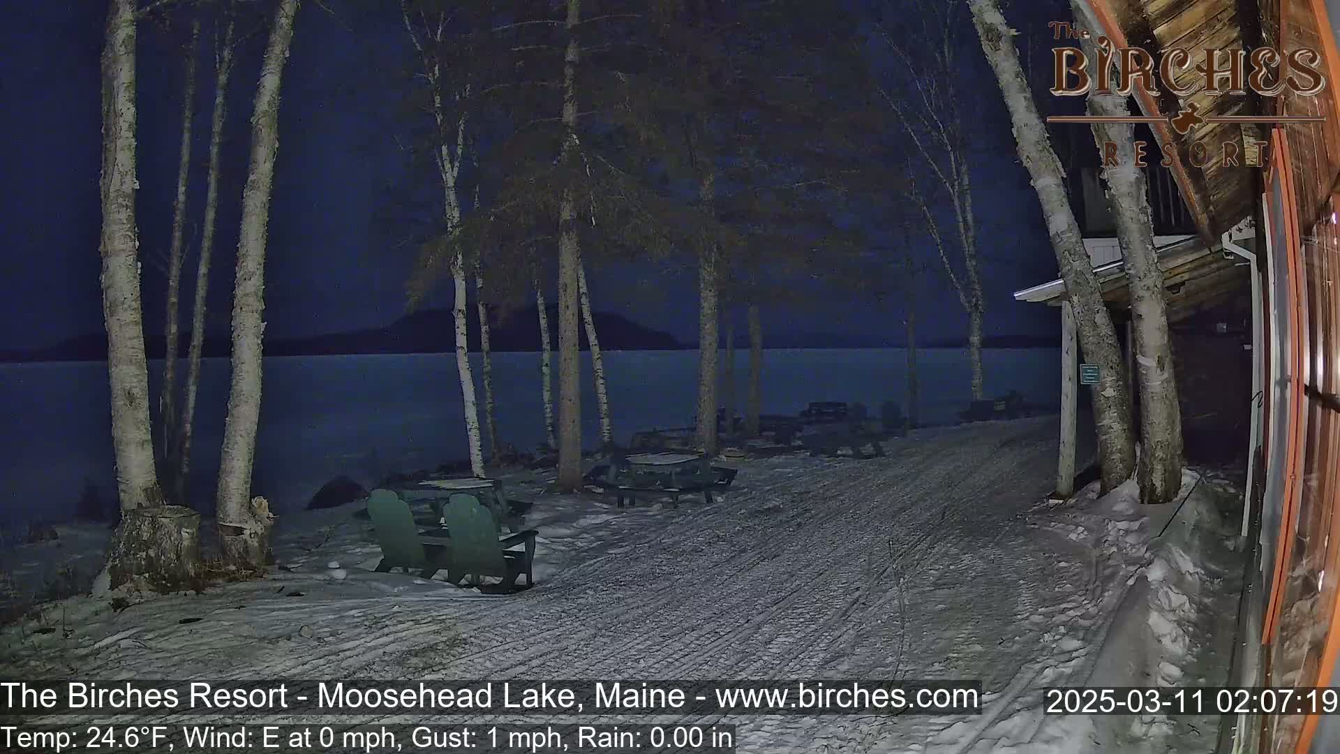 A snow-covered lakeside area at night, with picnic tables and chairs under birch trees, shows a calm lake in the background under a dark sky.