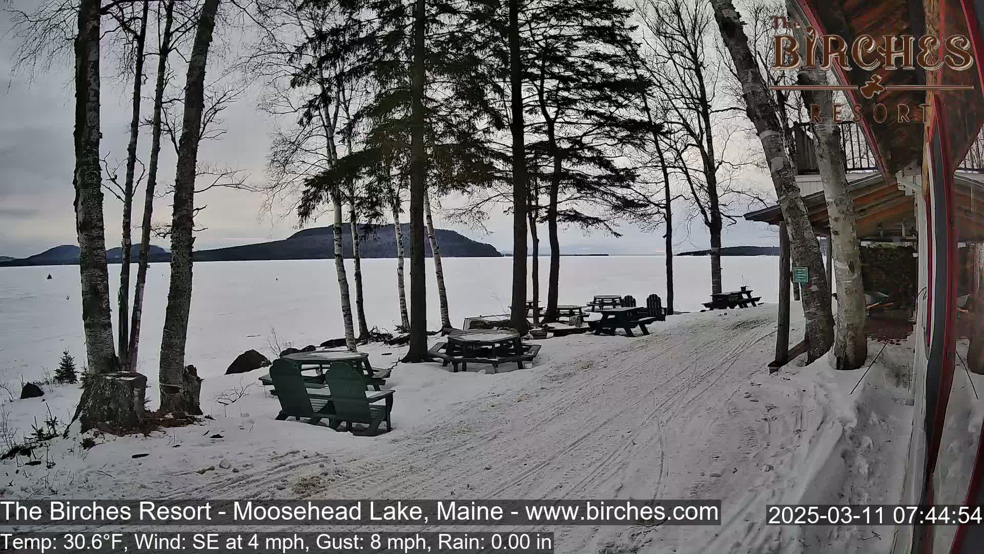A snow-covered lakeside area with picnic tables and Adirondack chairs under bare trees, overlooking a calm lake and distant hills under an overcast sky.