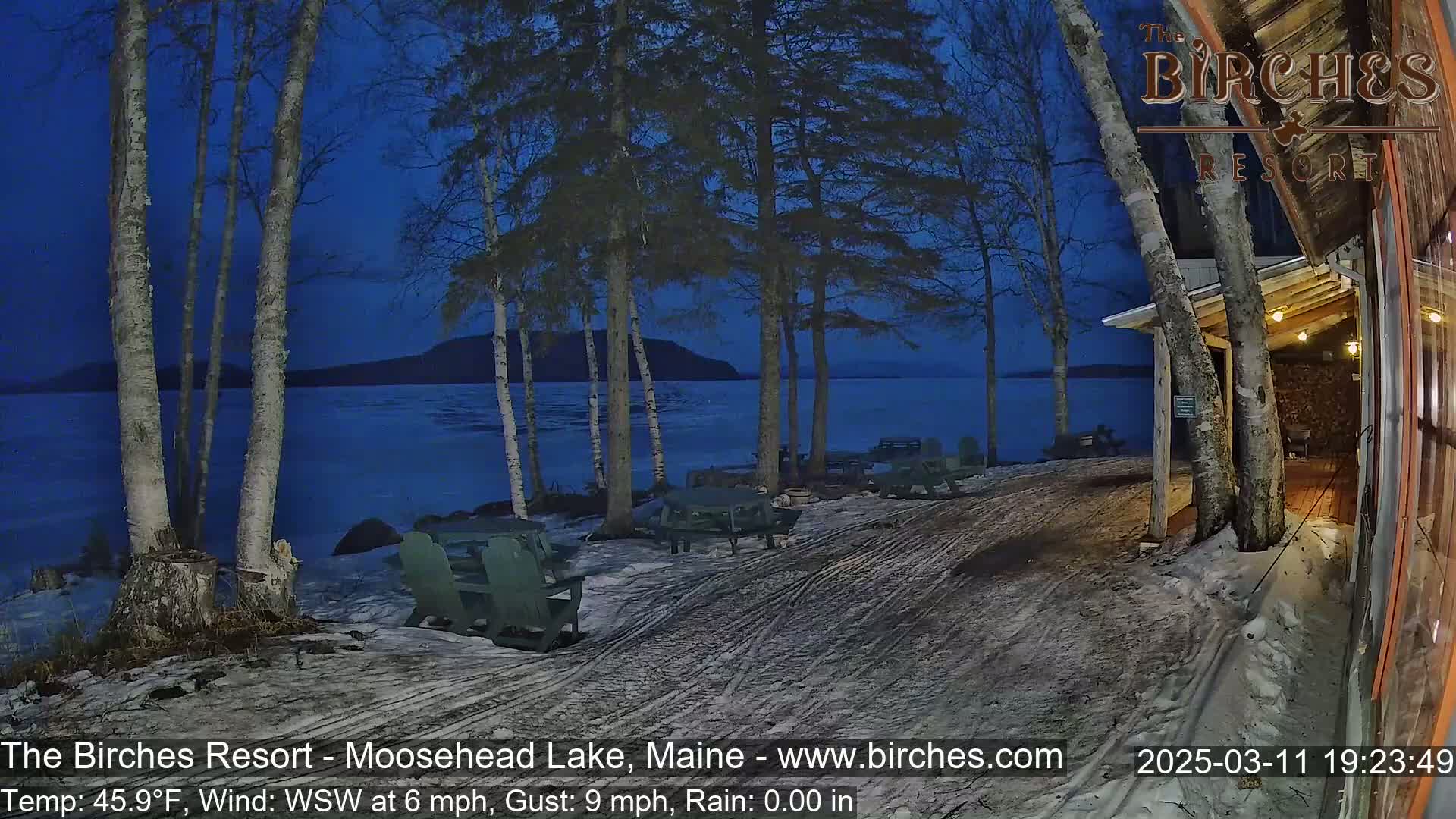 A snow-dusted lakeside patio with picnic tables sits under a twilight sky next to a building, overlooking a dark, partially frozen lake.
