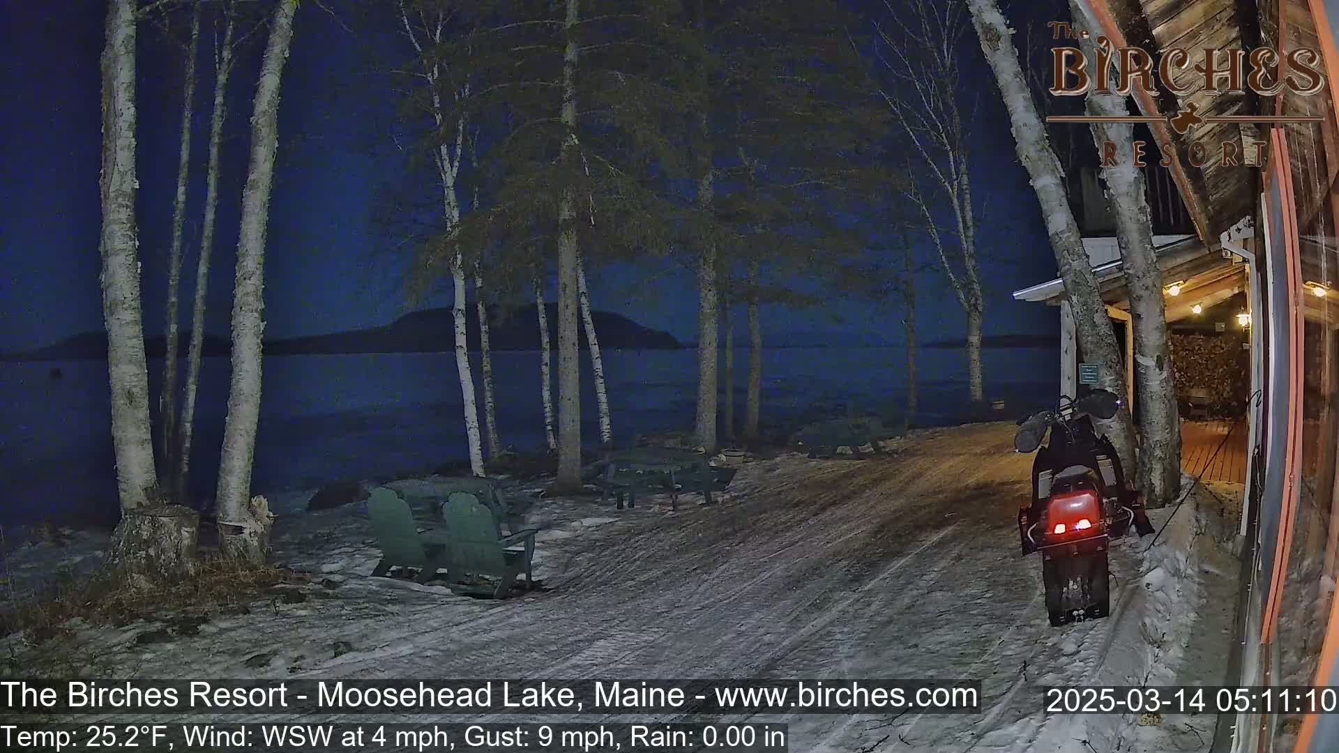 A snow-covered lakeside area at night under a dark sky, with birch trees, outdoor seating, and a snowmobile parked near a building, in rainy conditions.